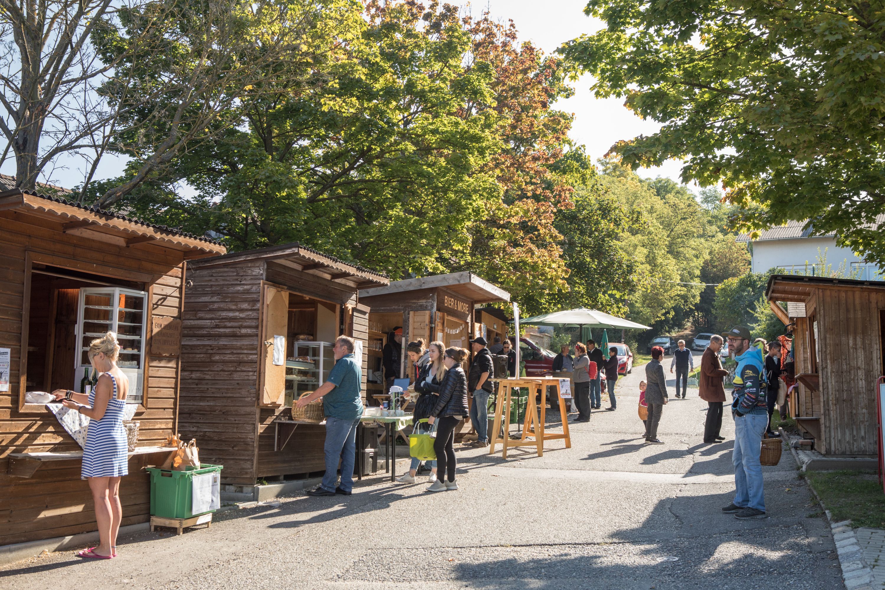 Menschen stehen an Holzhütten auf einem Markt im Freien bei sonnigem Wetter.