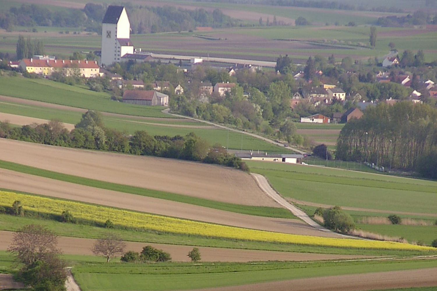 Landschaft mit Feldern und einem Dorf im Hintergrund, dominiert von einem großen weißen Gebäude mit dunklem Dach.