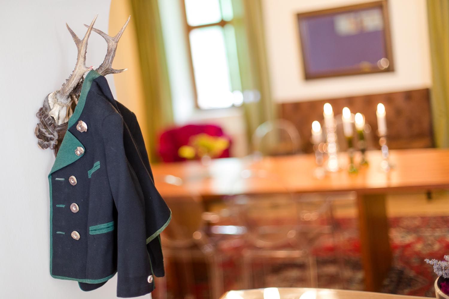 Ein traditioneller Janker hängt an einem Geweih an der Wand. Im Hintergrund ein Tisch mit Kerzenleuchtern und ein Fenster.