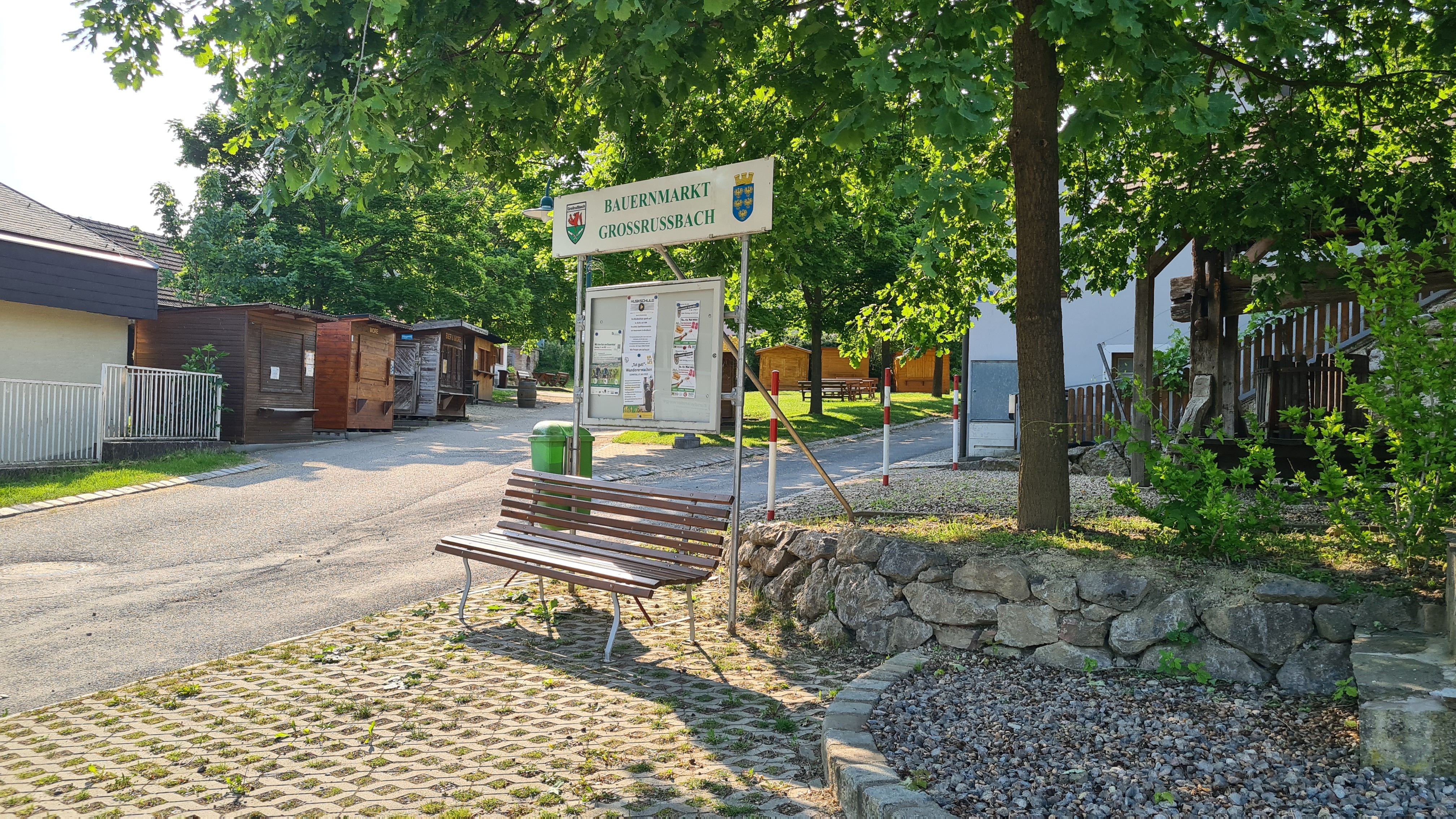 Vstup na farmářský trh v Großrußbachu s dřevěnými chatkami a lavičkami.