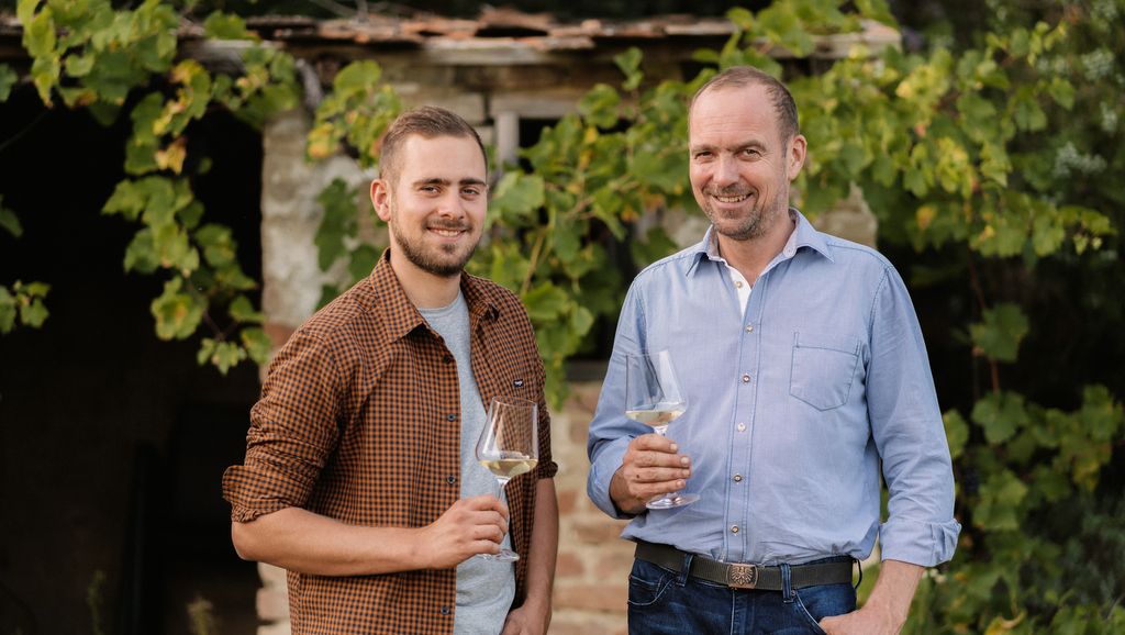 Zwei Männer stehen lächelnd mit Weingläsern vor einem Weinlaub bedeckten Gebäude.