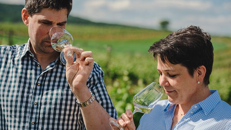 Ein Mann und eine Frau stehen in der Landschaft und riechen beide an einem Glas Weißwein.
