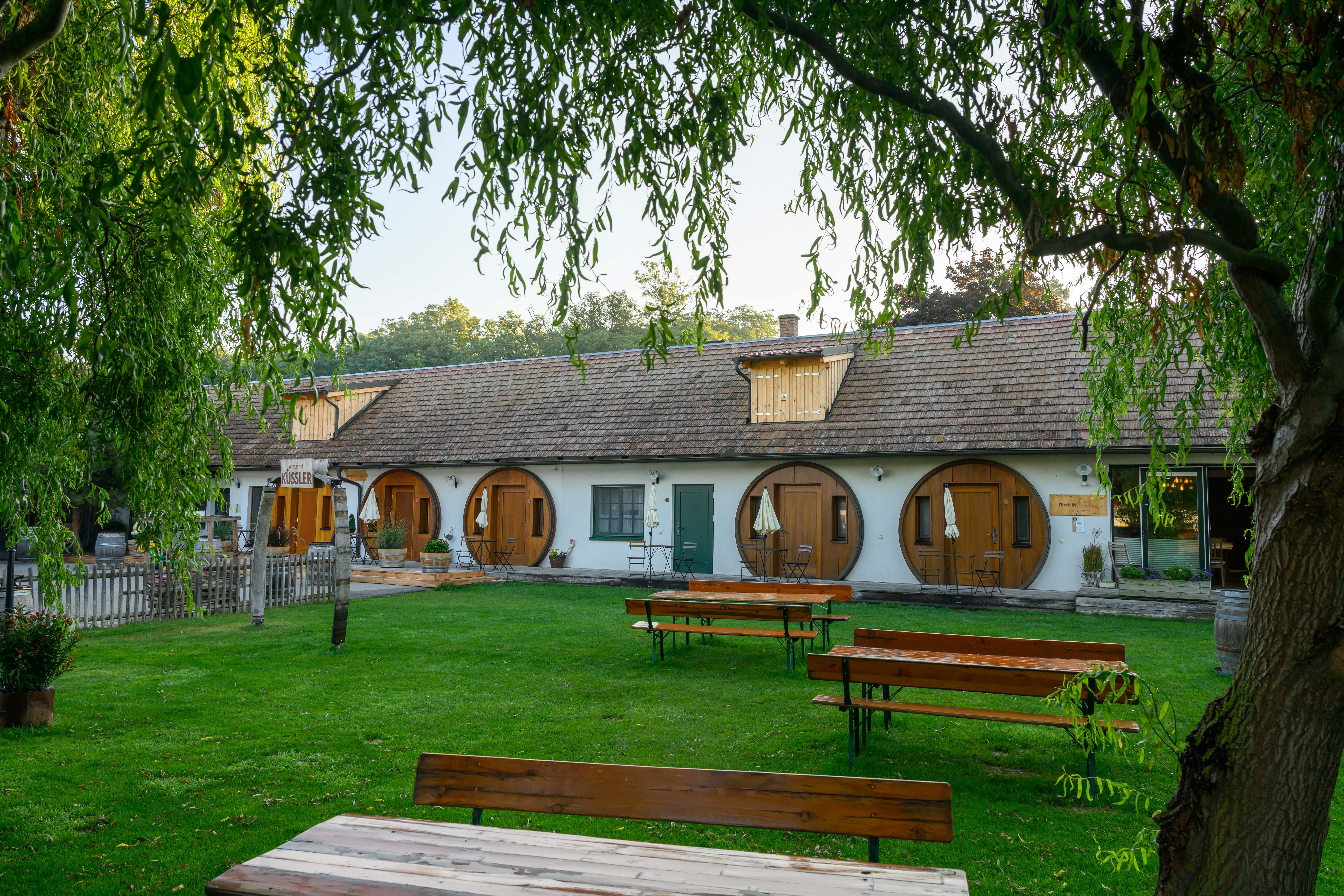 Winzerhof Küssler mit weißem Gebäude, runden Holztüren, grüner Wiese und Bänken unter überhängenden Bäumen.