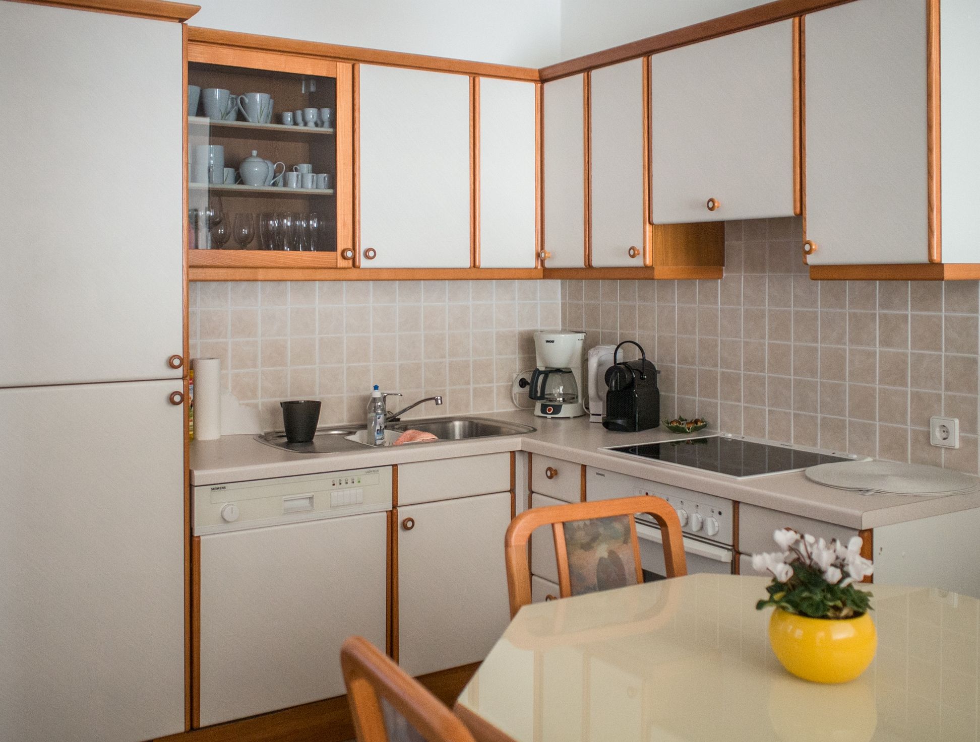 Moderne Küche mit weißen Schränken, Geschirrspüler, Kaffeemaschine und Herd. Gelber Blumentopf auf dem Tisch.