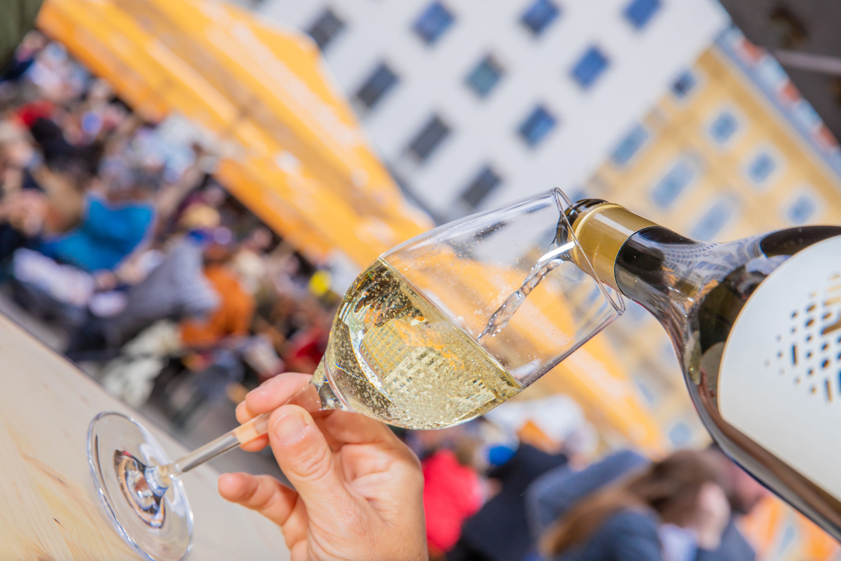 Im Vordergrund wird gerade ein Glas Weinviertler Sekt eingeschenkt, im Hintergrund sieht man Personen, welche Weinviertler Getränke und Kulinarik in einer Innenstadt genießen. 