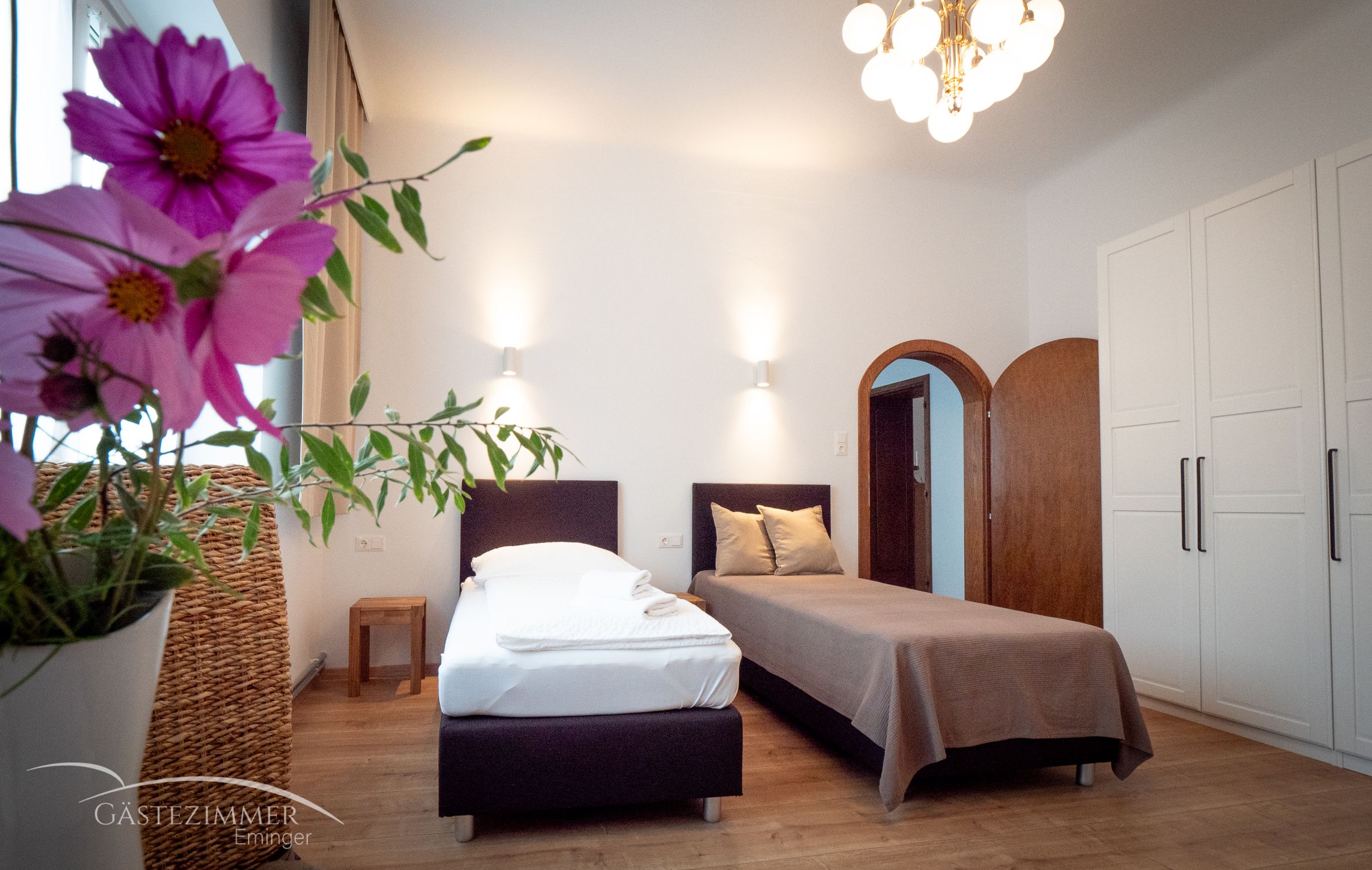 Ein gemütliches Zweibettzimmer mit Blumen im Vordergrund, zwei Betten, einem Schrank und einer Lampe an der Decke.