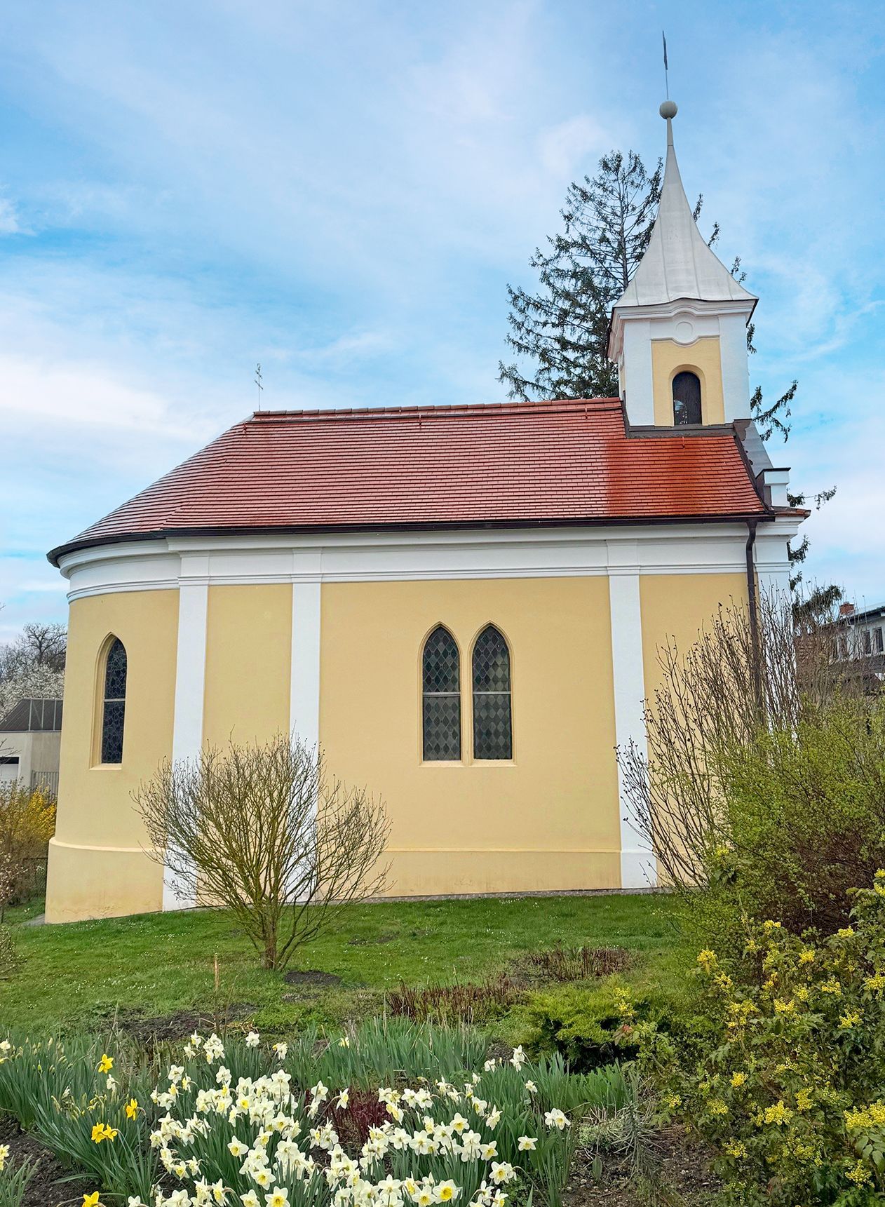 Eine kleine gelbe Kapelle mit rotem Dach und einem Turm, umgeben von blühenden Blumen und Büschen.
