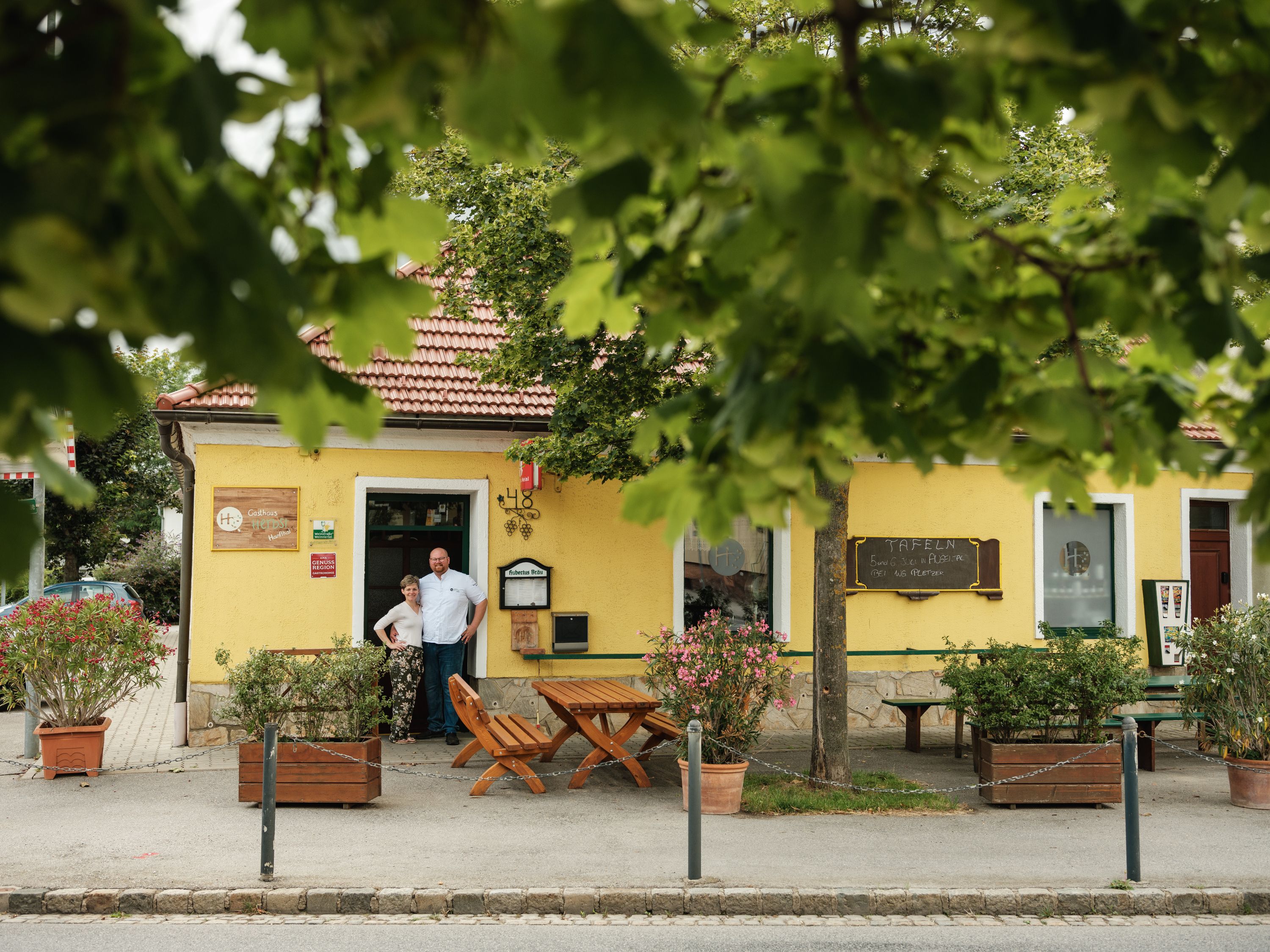 A yellow inn with a red roof, surrounded by trees. Two people are standing in front of the entrance.