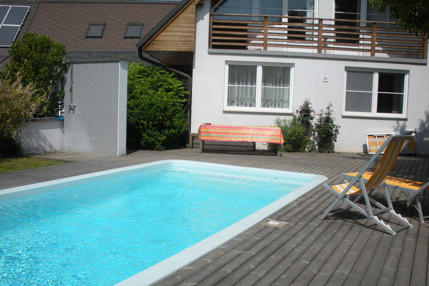 Ein privater Garten mit einem rechteckigen Schwimmbecken, umgeben von Holzterrasse, Liegestühlen und einem Haus im Hintergrund.
