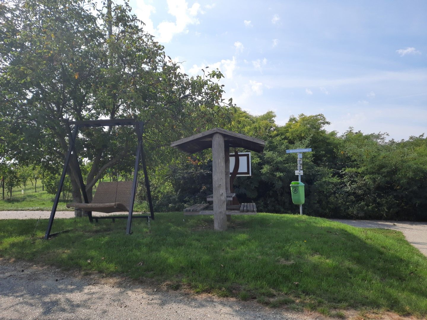 Ein Rastplatz mit einer Schaukel und einem überdachten Sitzbereich neben einem Baum und einem Mülleimer auf einer Wiese.