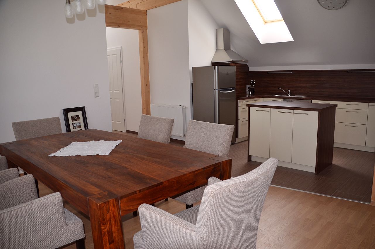 Modern dining area with wooden table and gray chairs, adjacent to a kitchen with island and skylight.