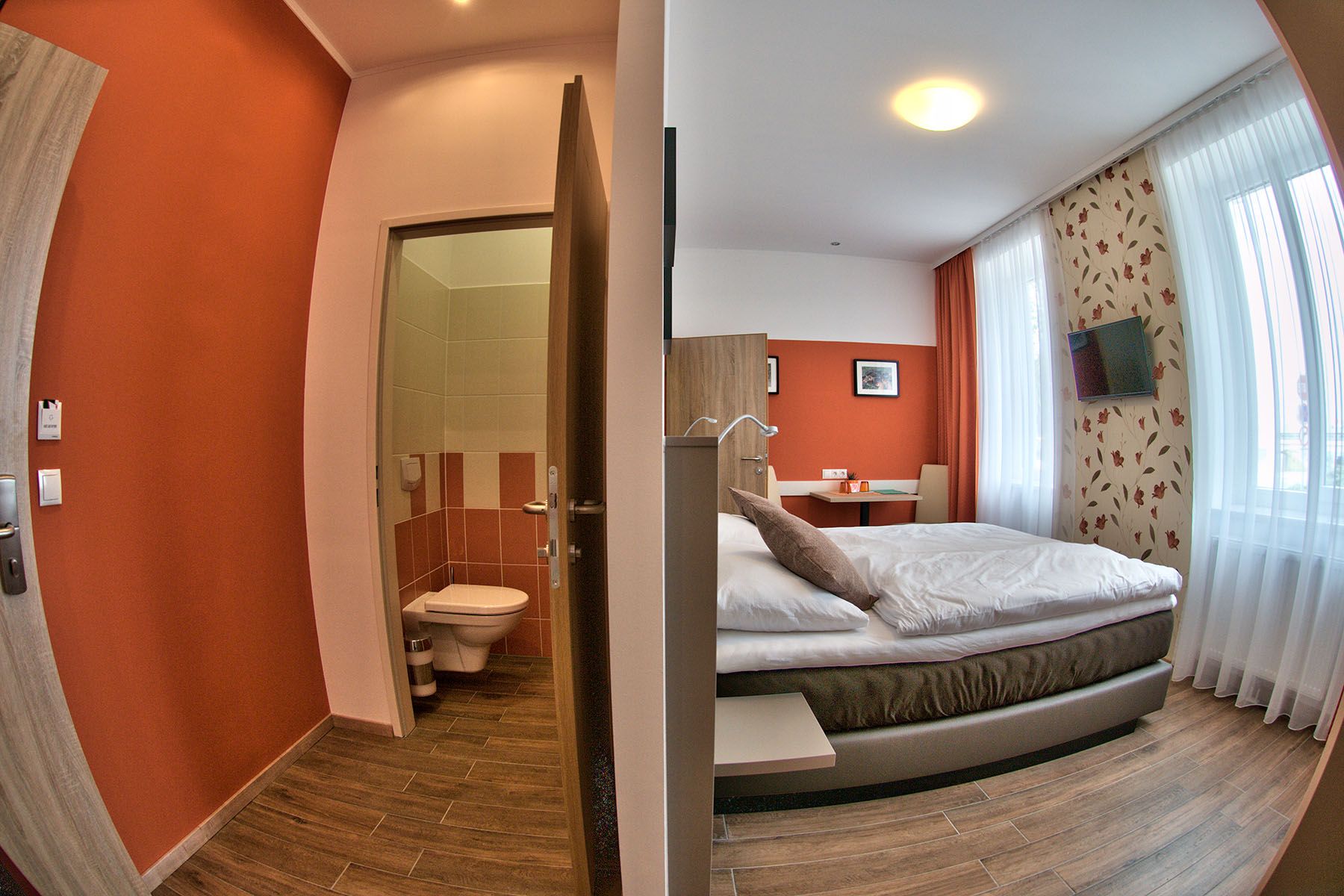 Ein modernes Gästezimmer mit Doppelbett, Schreibtisch und Fernseher. Daneben ein kleines Badezimmer mit Toilette.