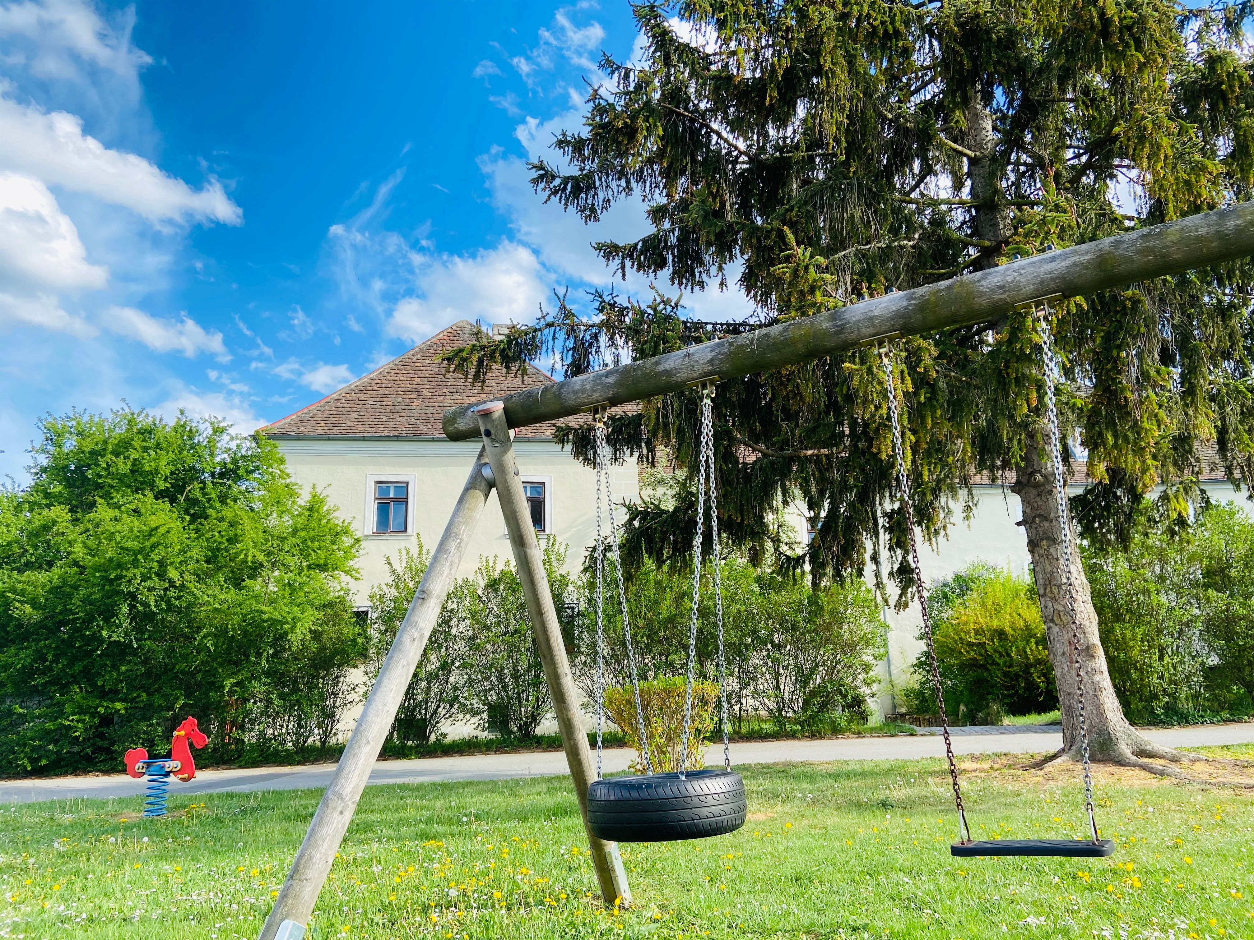 Spielplatz mit Schaukel und Wippe vor einem Gebäude mit rotem Dach.