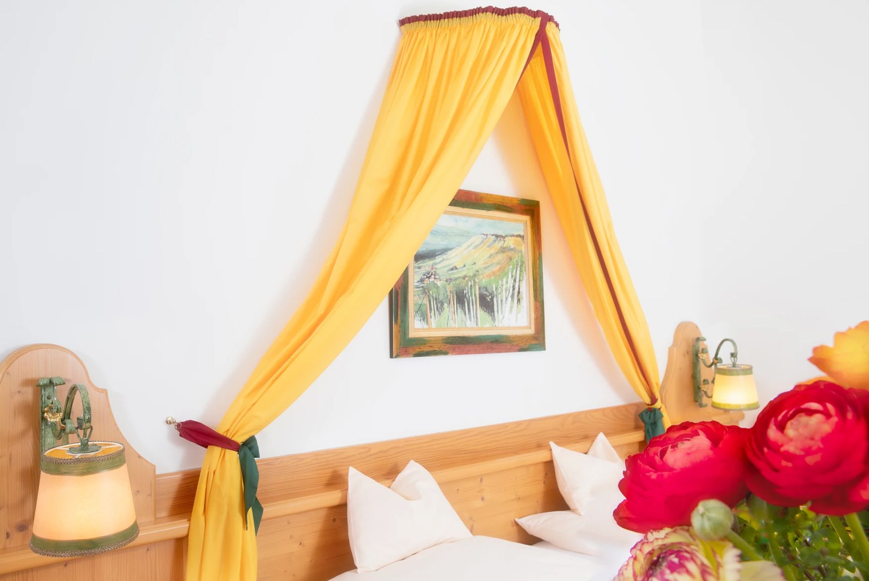 Ein gemütliches Doppelzimmer mit gelbem Baldachin, Holzbett, Wandlampen und einem Blumenstrauß im Vordergrund.