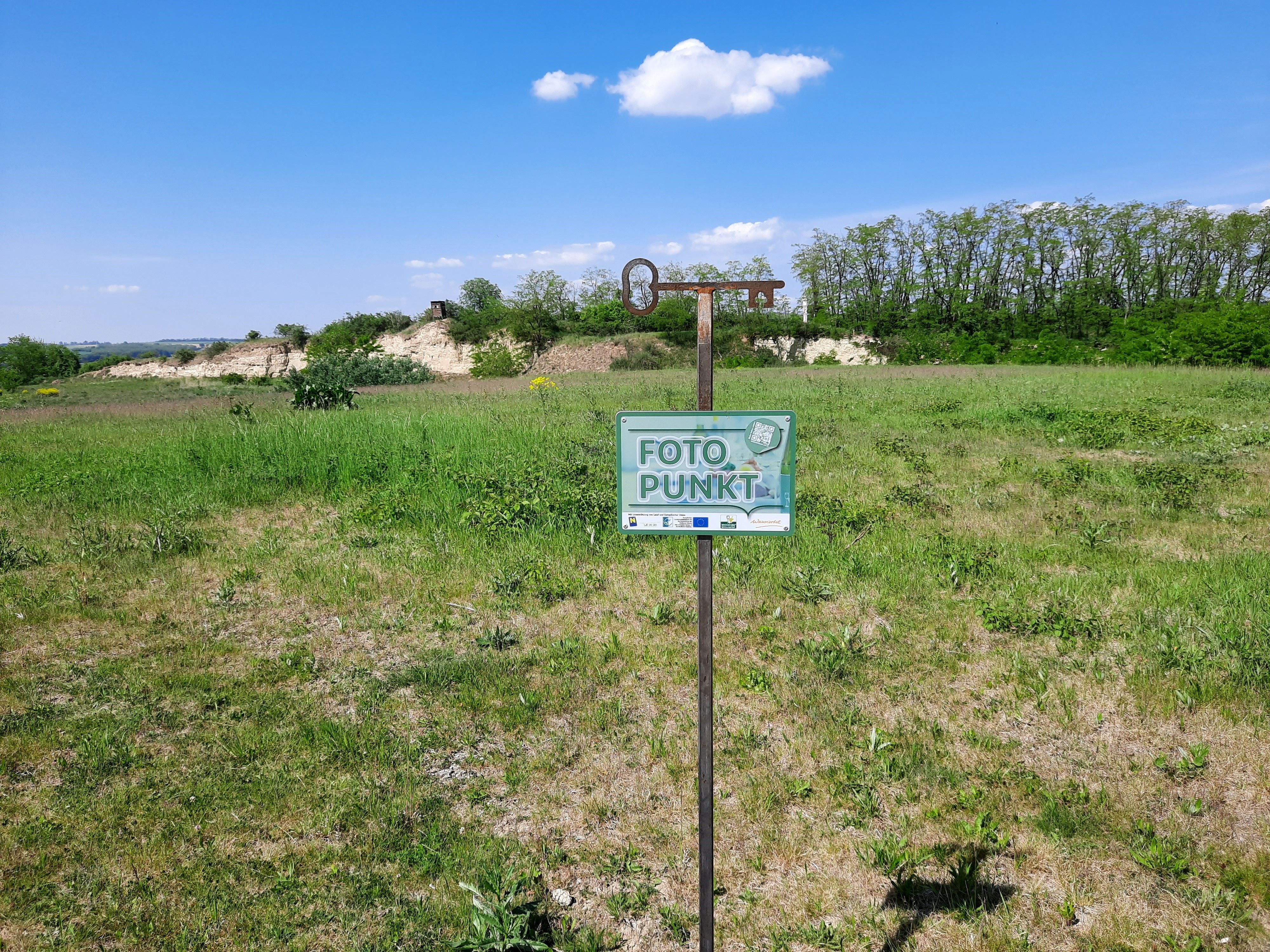 Ein Schild mit der Aufschrift 'Foto Punkt' steht auf einer grünen Wiese unter blauem Himmel.