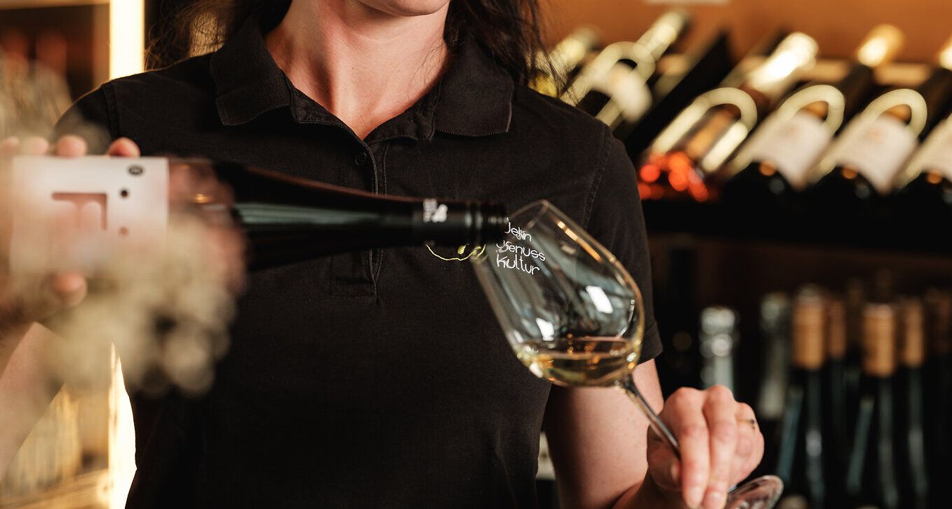 In einer einladenden Vinothek wird mit Hingabe ein edler Wein eingeschenkt. Die warmen Holztöne und die sorgfältig ausgewählten Flaschen schaffen eine gemütliche Atmosphäre, die zum Verweilen einlädt. Hier erleben Besucher die Leidenschaft für Wein und die kulinarischen Köstlichkeiten der Region.