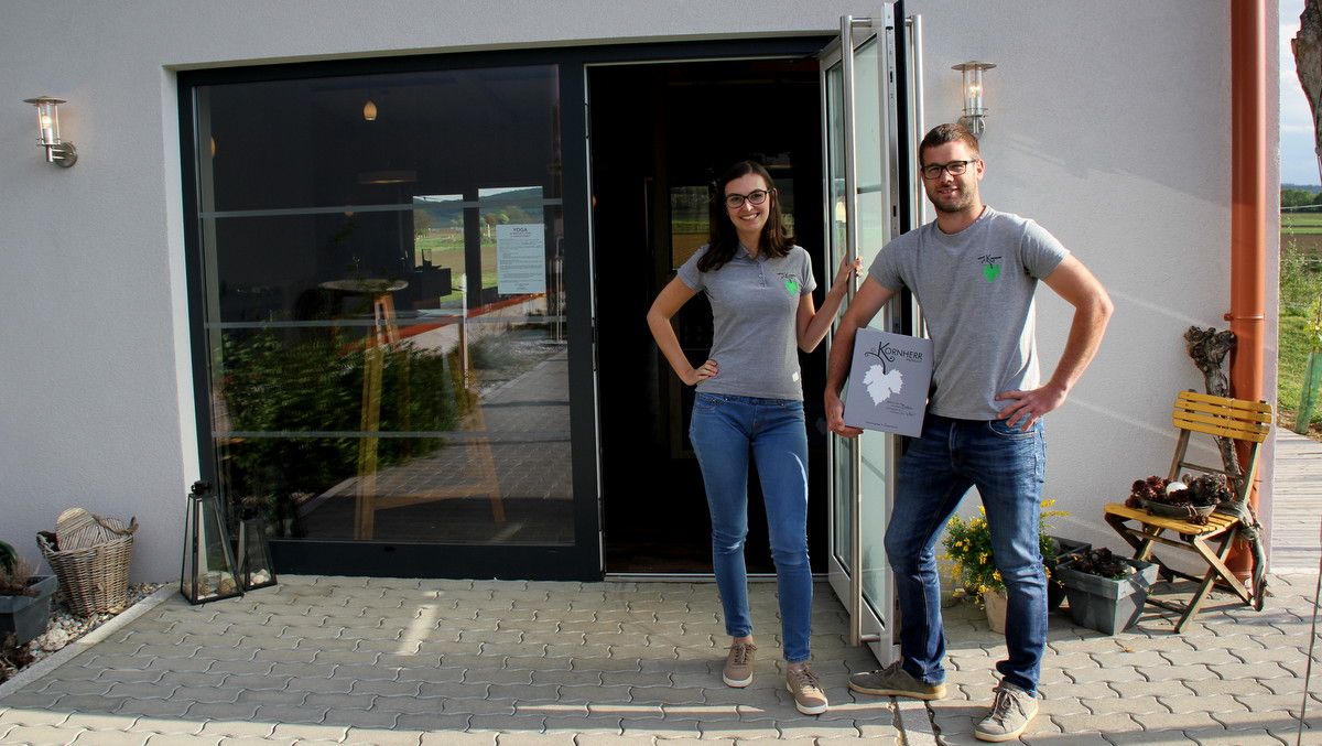 Zwei Personen stehen vor einem Gebäude mit Glastür, beide tragen graue T-Shirts und Jeans. Eine Person hält ein Schild mit der Aufschrift 'Kornherr'.