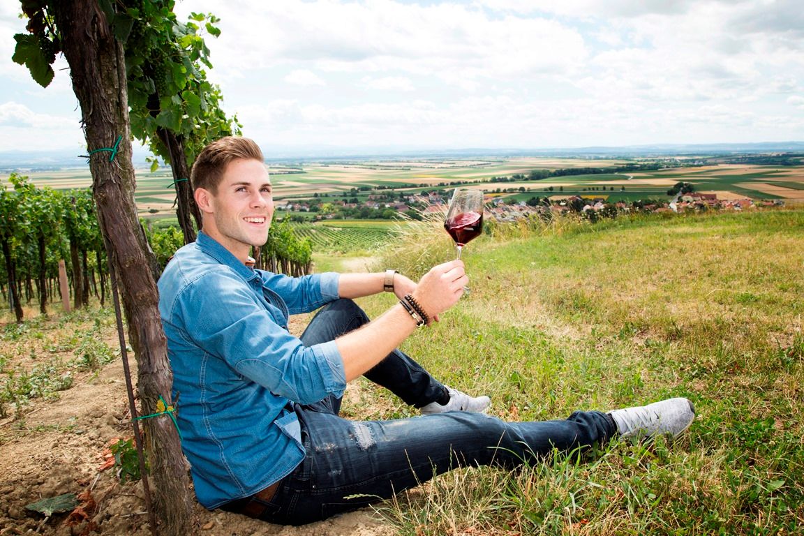 Ein Mann sitzt in einem Weinberg und hält ein Glas Rotwein. Im Hintergrund ist eine weite Landschaft zu sehen.
