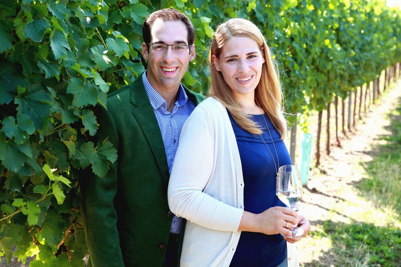 A smiling couple stands in a vineyard, the woman holding a glass of wine.