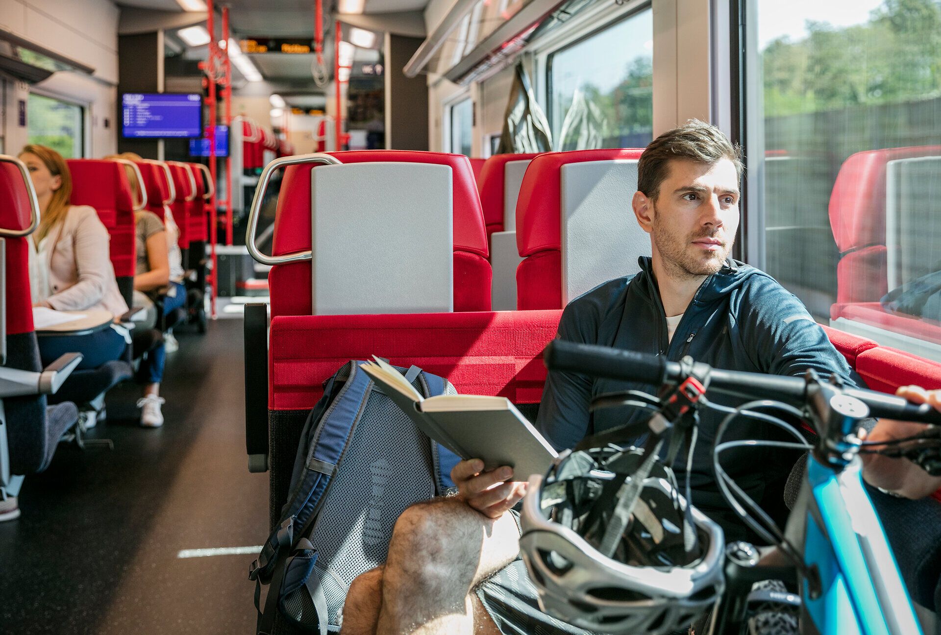 Eine entspannte Zugfahrt durch die malerische Landschaft bietet die perfekte Gelegenheit, die Schönheit der Natur zu genießen. Der Reisende mit seinem Fahrrad und einem Buch in der Hand lässt sich von der vorbeiziehenden Kulisse inspirieren und träumt von neuen Abenteuern in den Bergen.