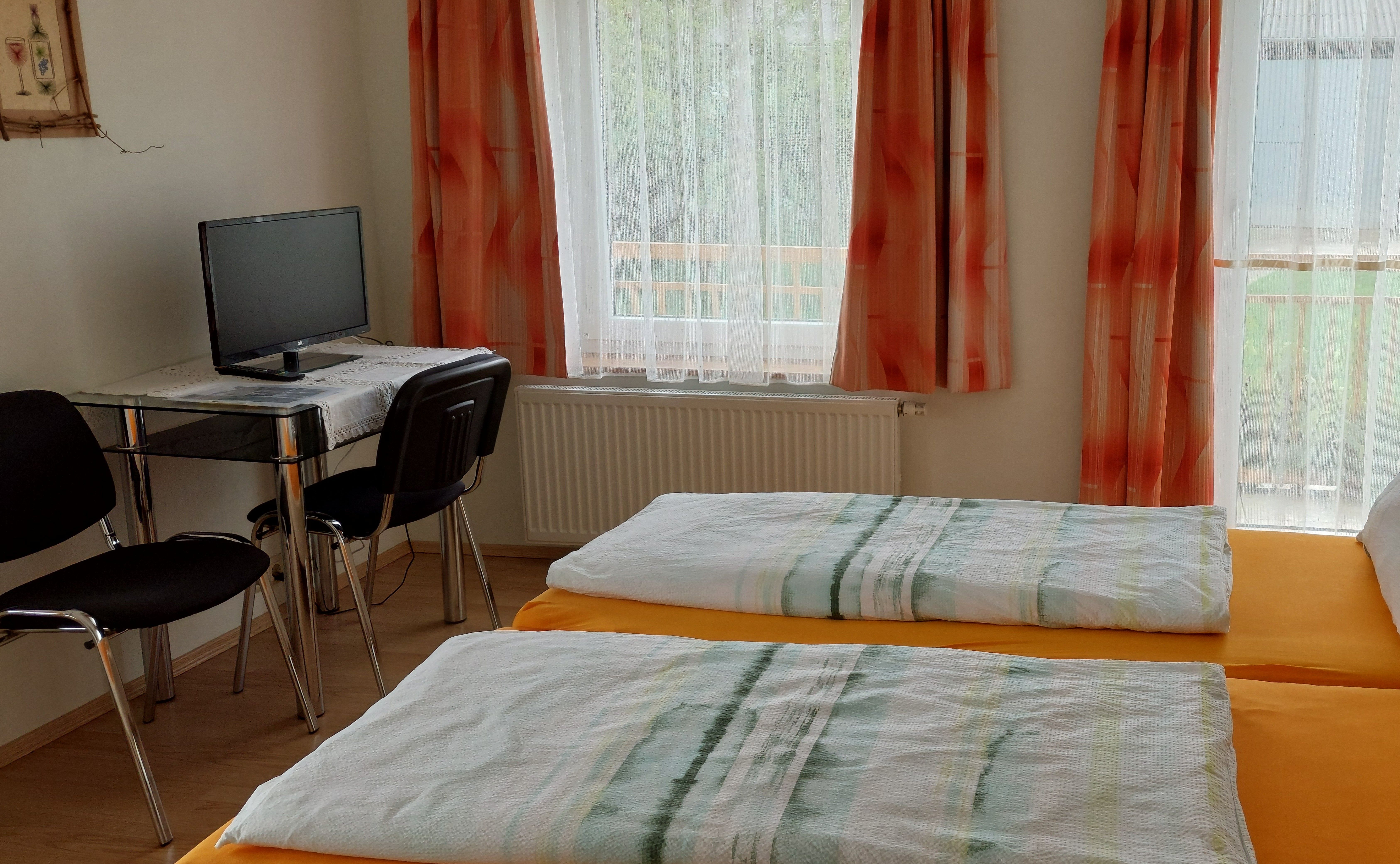 Ein einfach eingerichtetes Zimmer mit zwei Einzelbetten, einem kleinen Tisch mit Stuhl und einem Fernseher. Die Fenster sind mit orangefarbenen Vorhängen dekoriert.