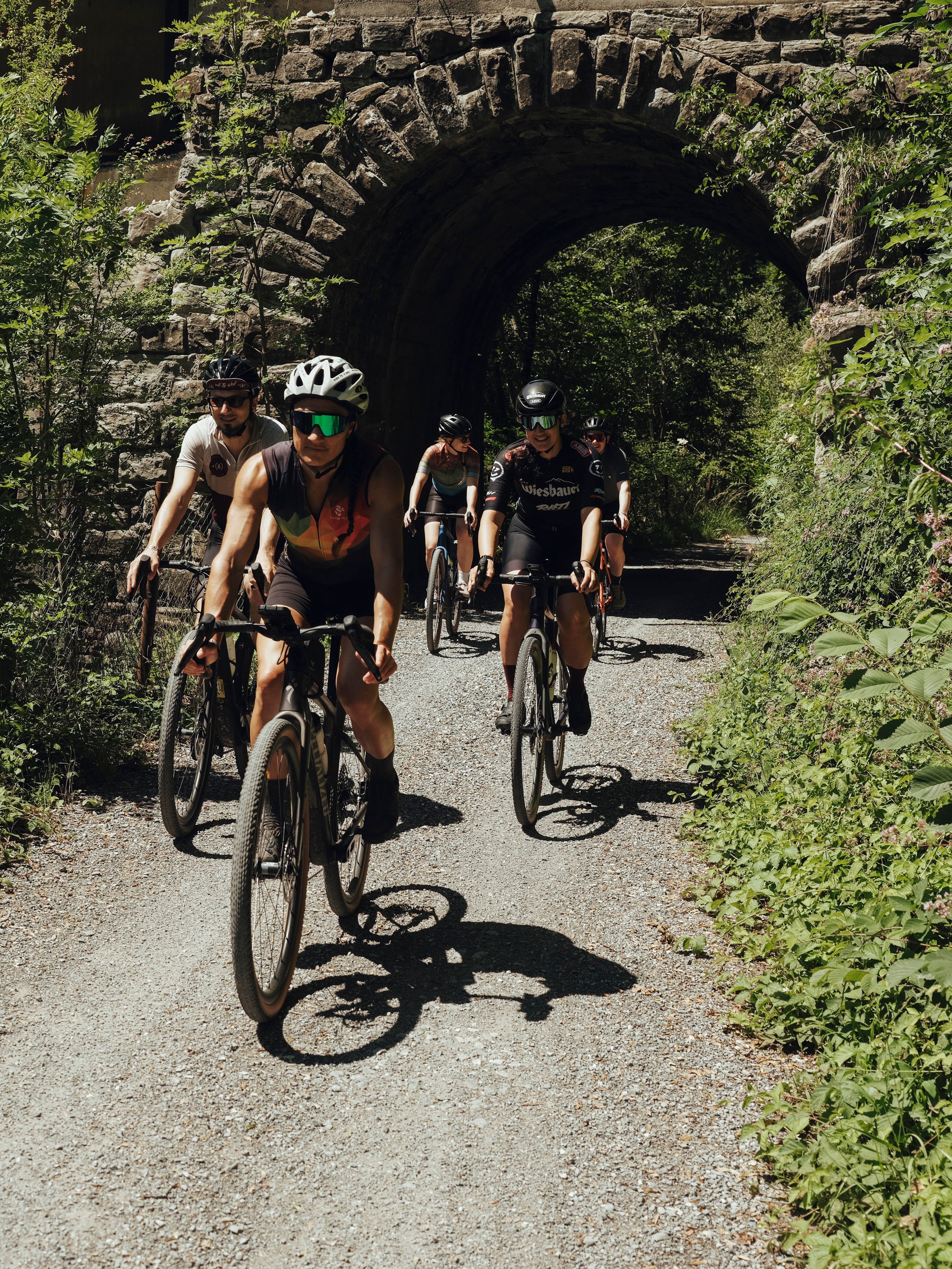 Gruppe von Radfahrern fährt auf einem Schotterweg unter einem steinernen Torbogen hindurch.