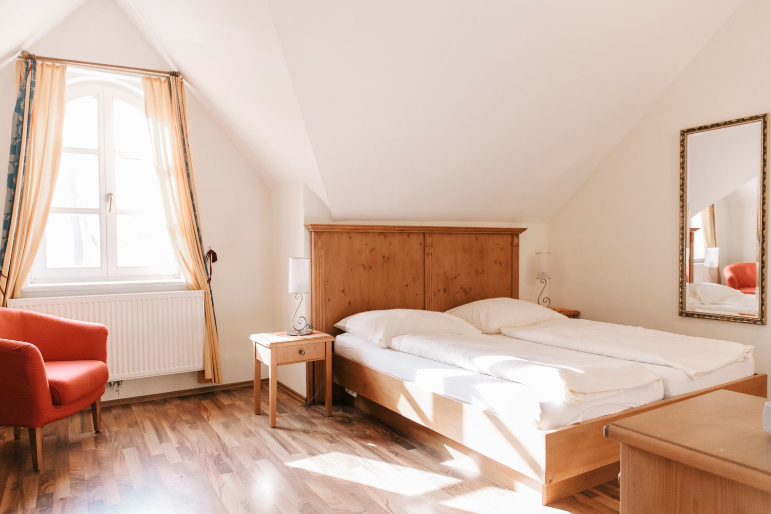 Helles Gästezimmer mit Doppelbett, rotem Sessel und großem Fenster mit Vorhängen.