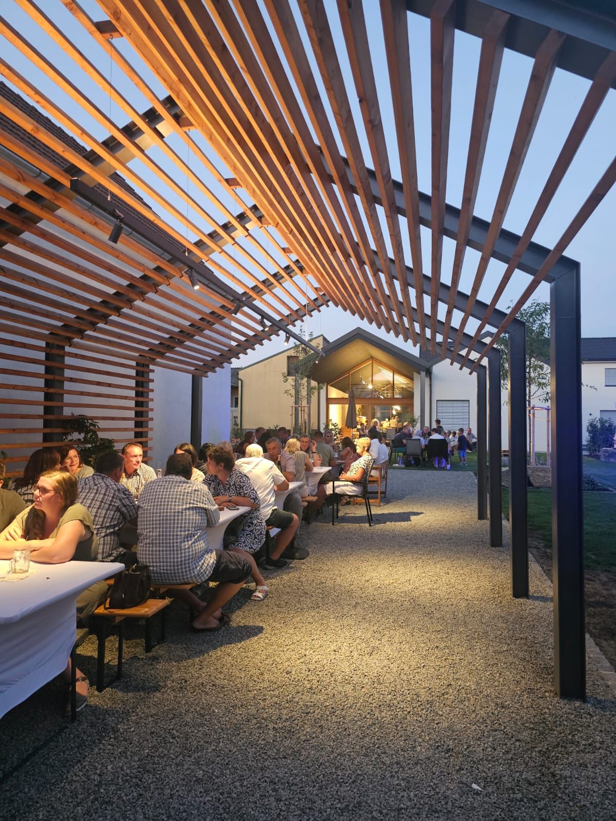 Menschen sitzen an langen Tischen unter einem Holzpergola bei Abenddämmerung.