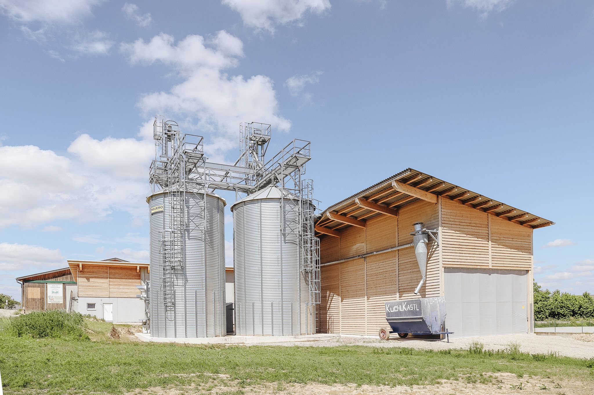 Betriebs-Ansicht mit Holzgebäude, Silo und umliegenden Feldern.