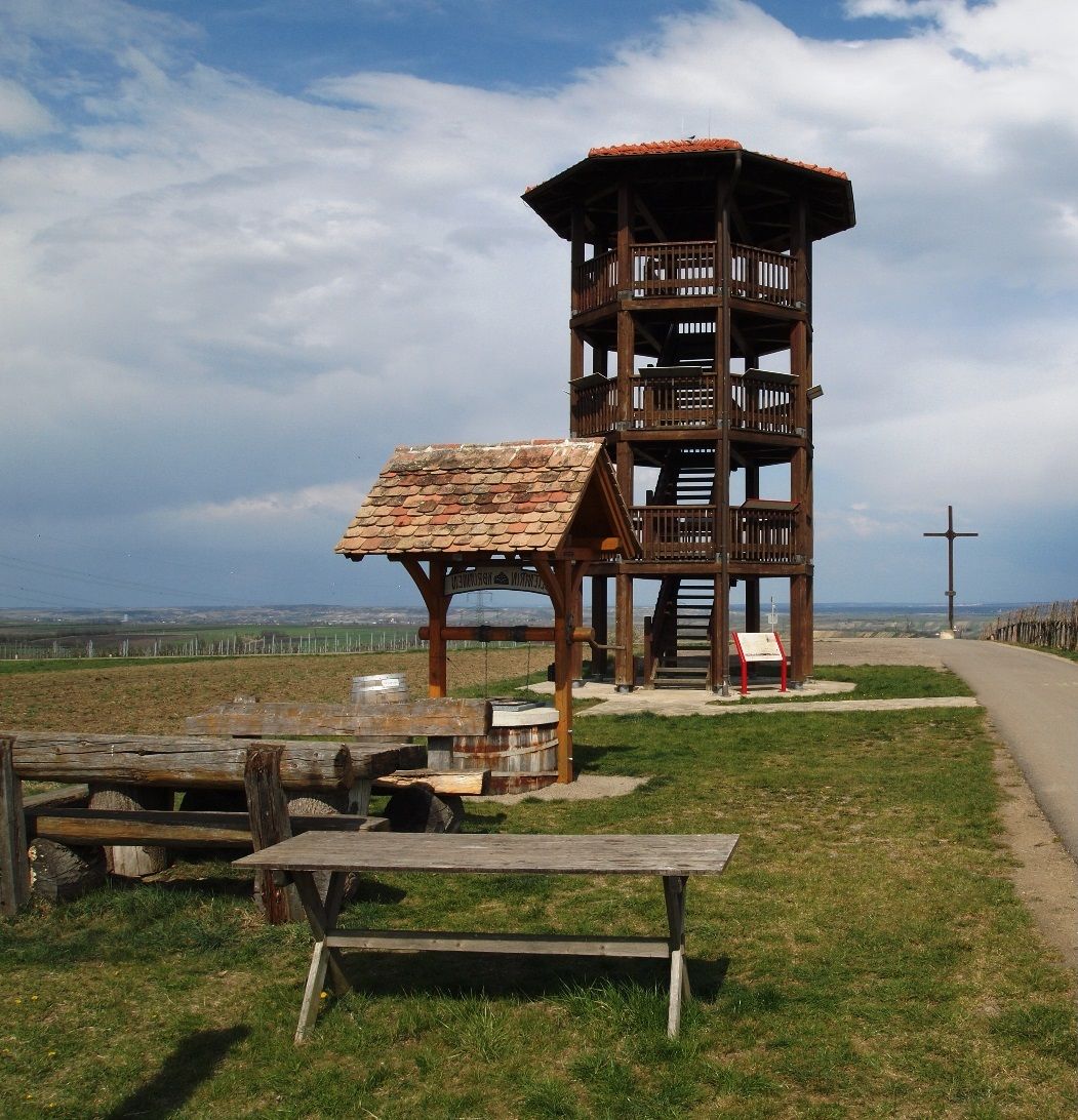 Aussichtsturm aus Holz auf einer Wiese mit Sitzbänken und einem Kreuz im Hintergrund.