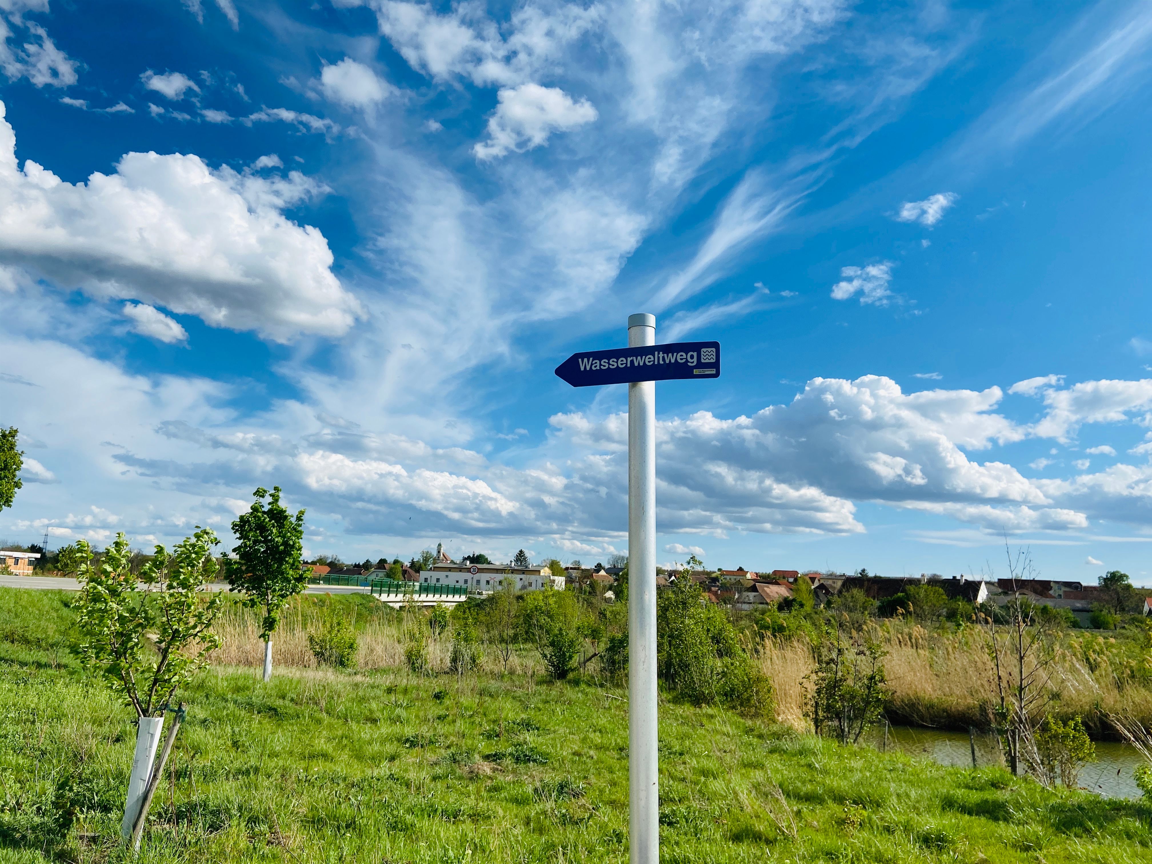 Ein Wegweiser mit der Aufschrift 'Wasserweltweg' steht auf einer grünen Wiese unter einem blauen Himmel mit weißen Wolken.