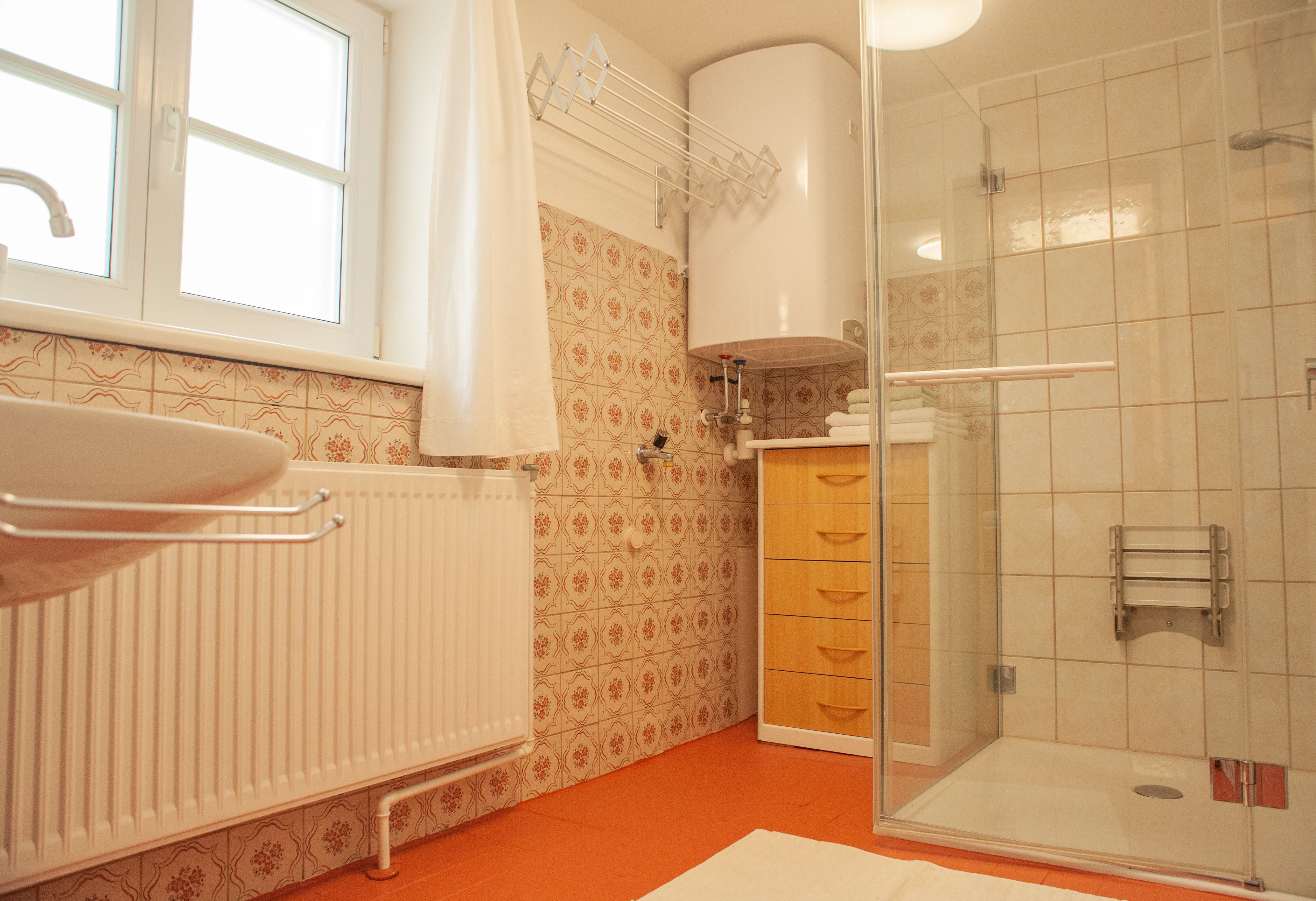 Kúpeľňa so sprchovacím kútom, umývadlom, radiátorom a drevenou skrinkou. Steny sú obložené vzorovanými dlaždicami.