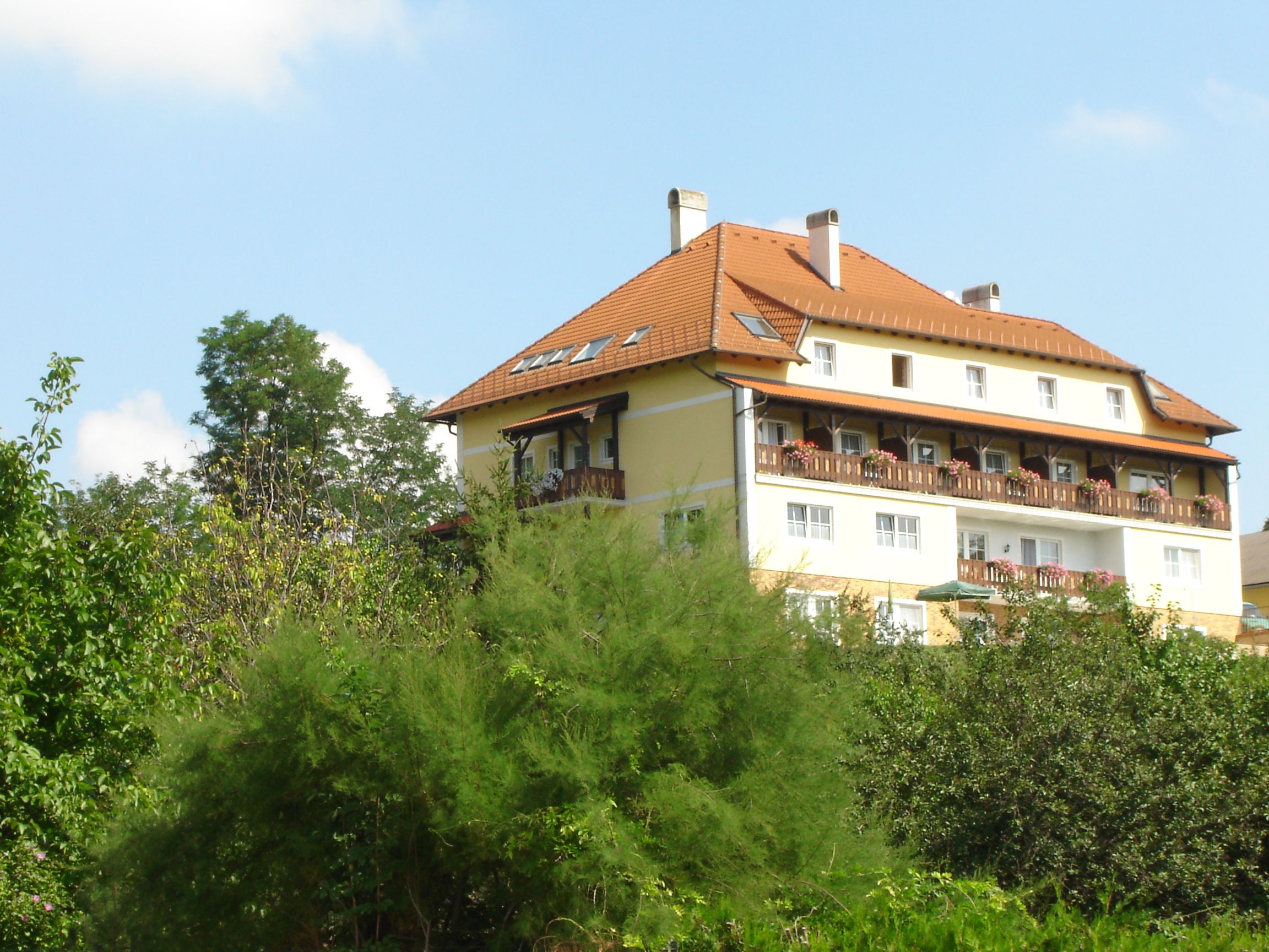 Ein großes, mehrstöckiges Gebäude mit rotem Dach und Balkonen, umgeben von Bäumen und Pflanzen.