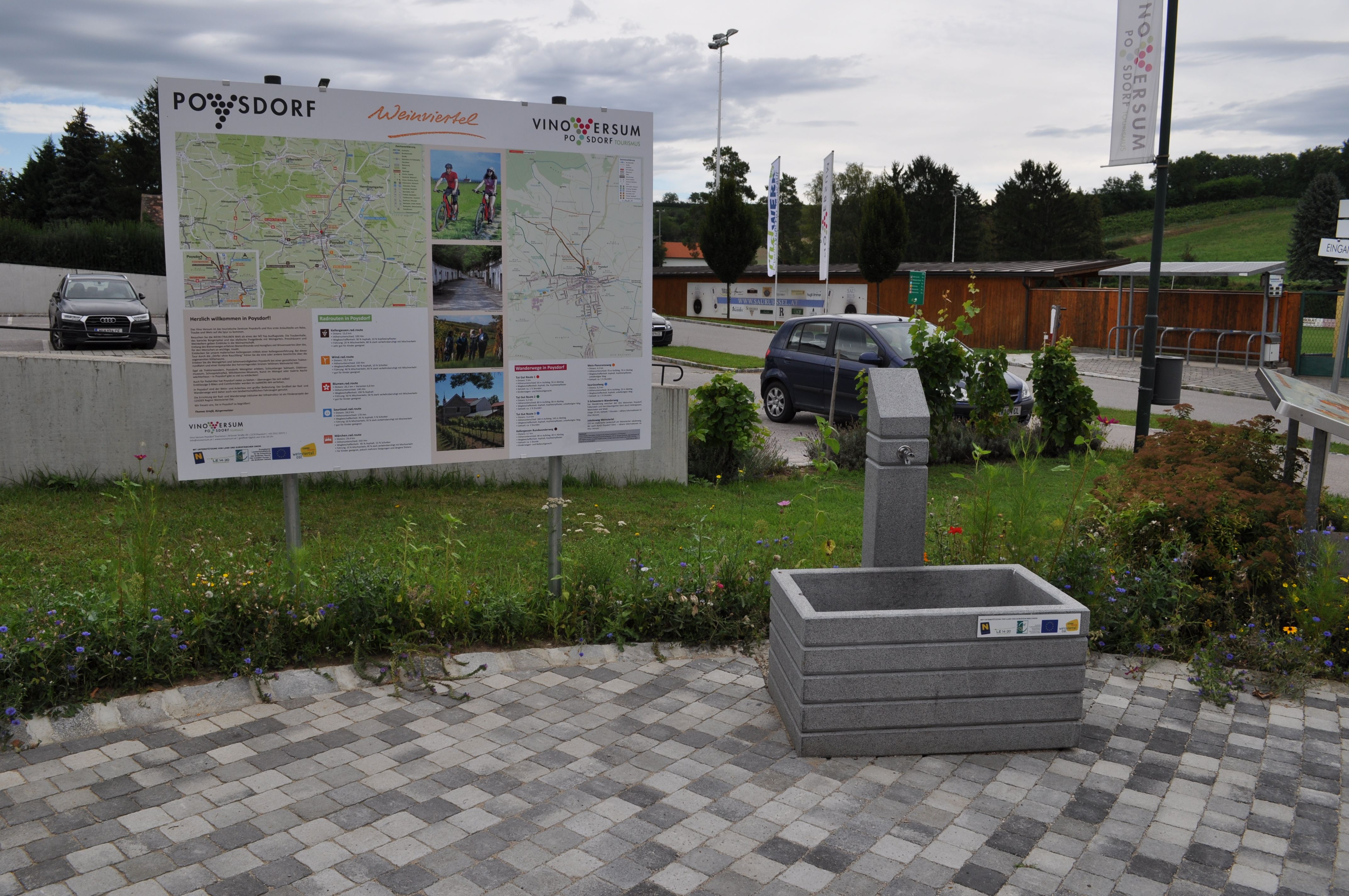 A drinking fountain next to an information board in Poysdorf, surrounded by flowers and parked cars.