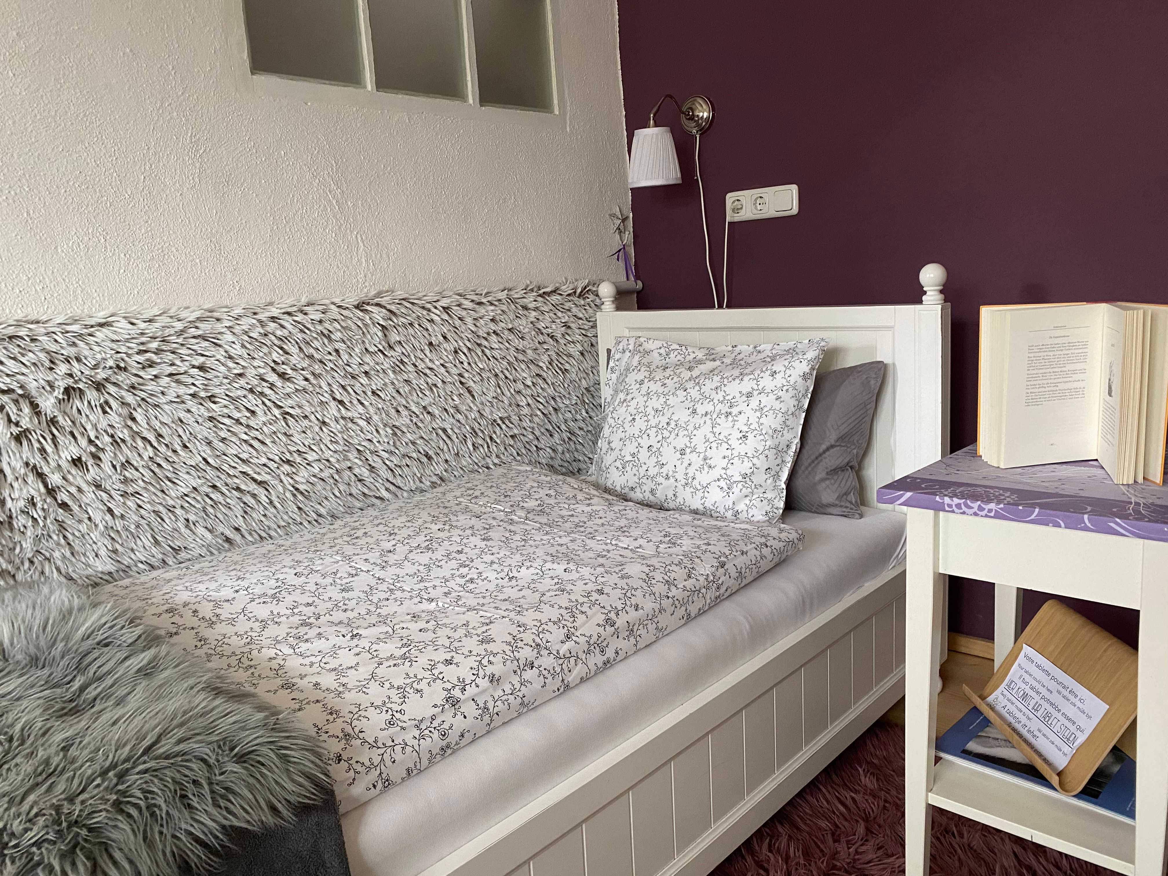 Gemütliches Schlafzimmer mit Einzelbett, lila Wand, Nachttisch mit Buch und Lampe.