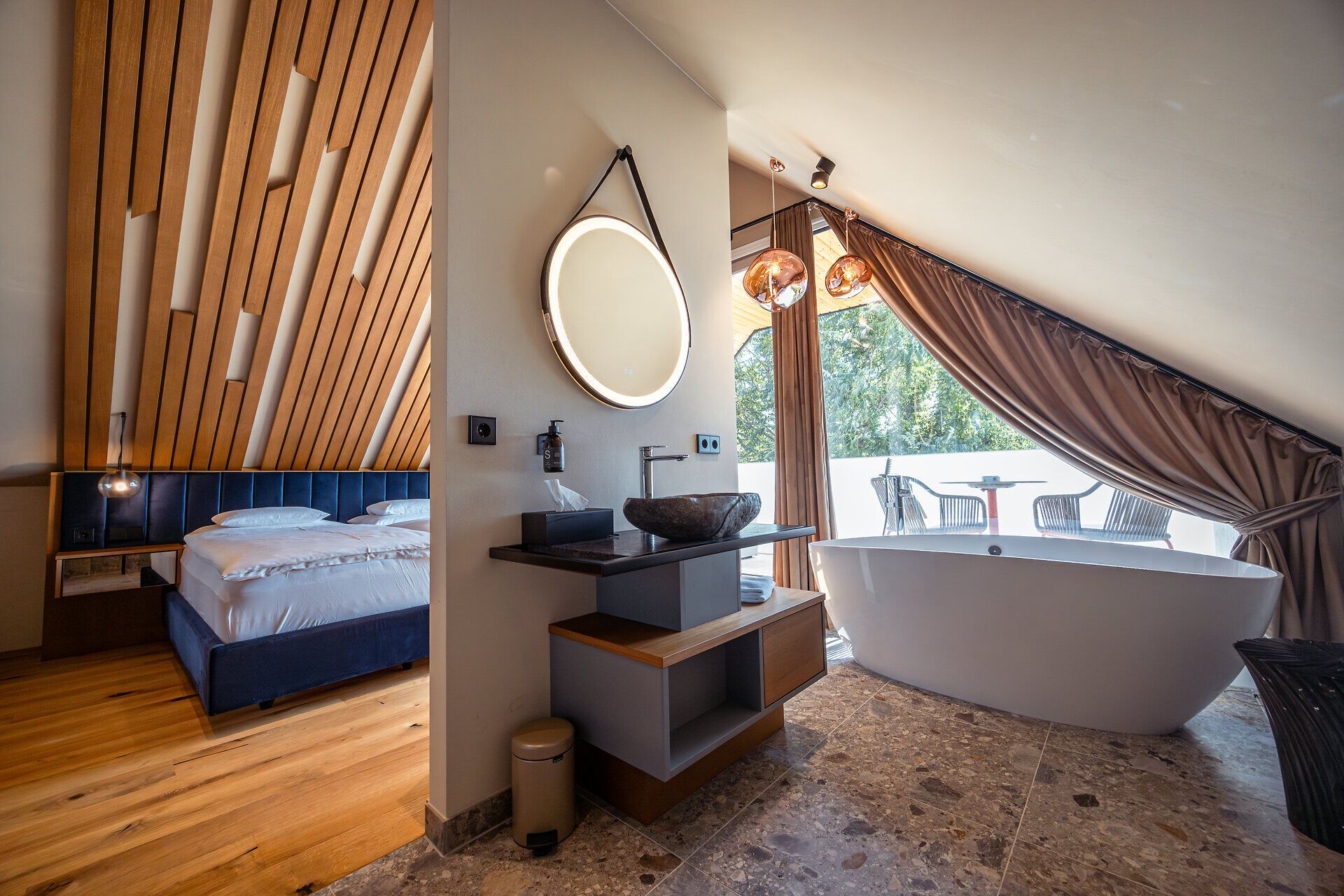 In einem stilvollen Raum mit modernem Design und warmen Holzelementen lädt eine freistehende Badewanne zum Entspannen ein. Der Blick auf die umliegenden Weinberge und die sanften Hügel schafft eine harmonische Atmosphäre, die zum Verweilen einlädt.