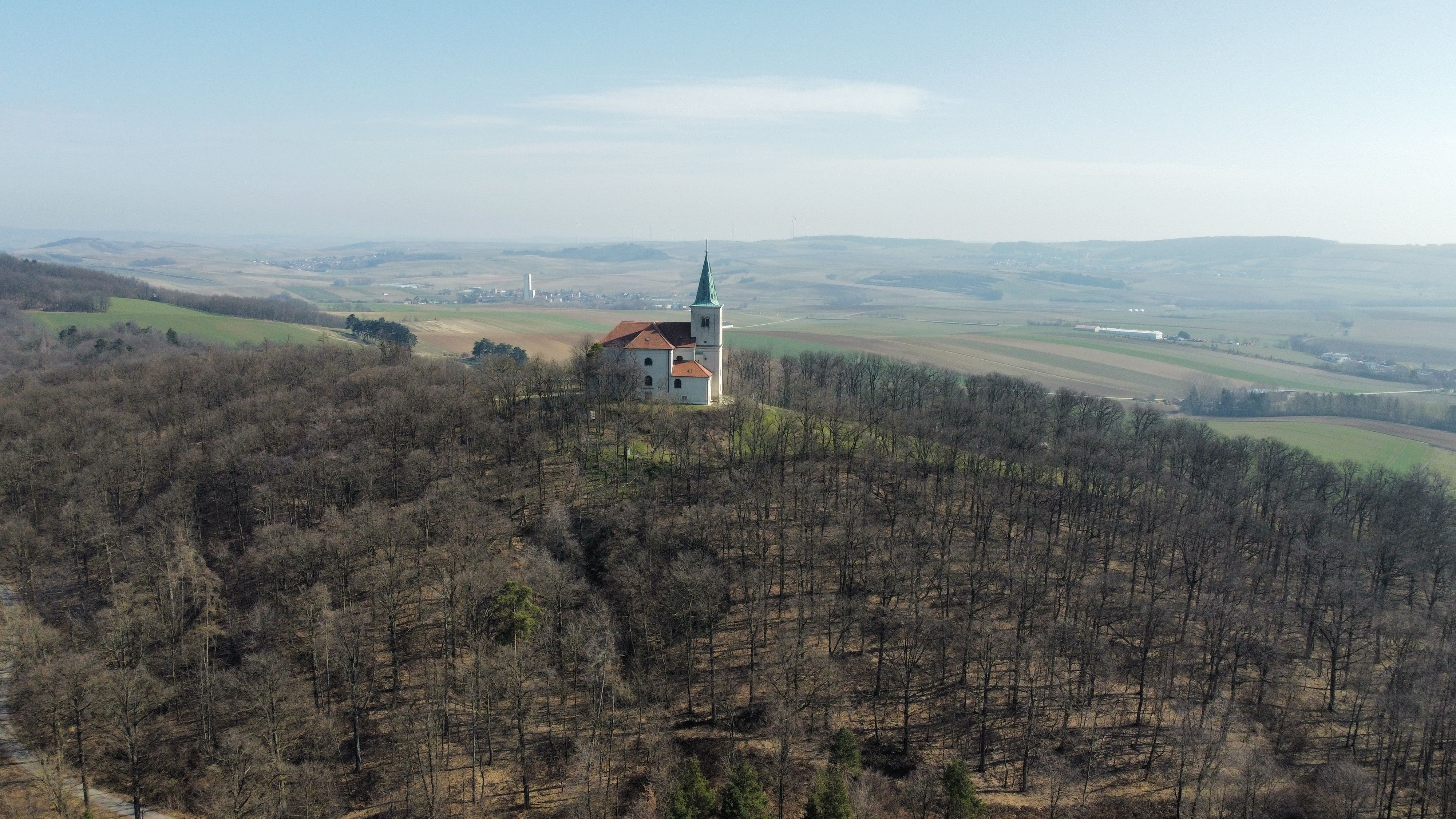 Luftaufnahme der Kirche Karnabrunn auf einem bewaldeten Hügel mit umliegender Landschaft.