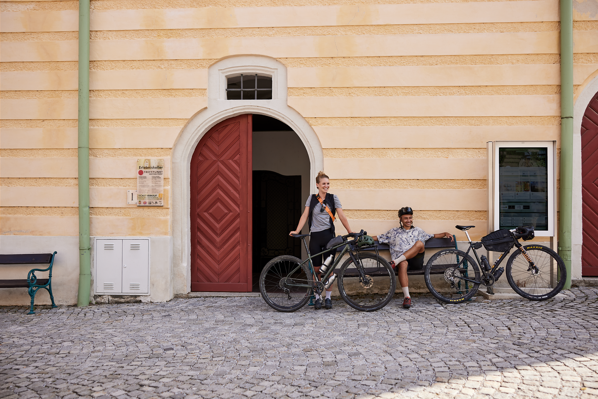 Zwei Radfahrer genießen eine wohlverdiente Pause vor einer charmanten, gelb gestrichenen Wand. Die Fahrräder stehen bereit für das nächste Abenteuer, während die Sonne sanft auf die Kopfsteinpflasterstraße scheint. Umgeben von der malerischen Landschaft des Weinviertels, lädt dieser Ort dazu ein, die Schönheit der Natur zu erkunden.