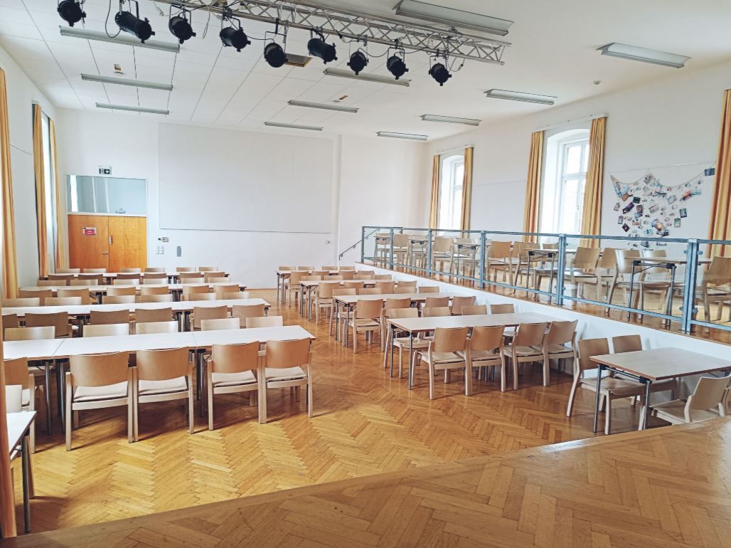 Prázdna sála s drevenou podlahou, stolmi a stoličkami, pódiom a osvetlením na strope.