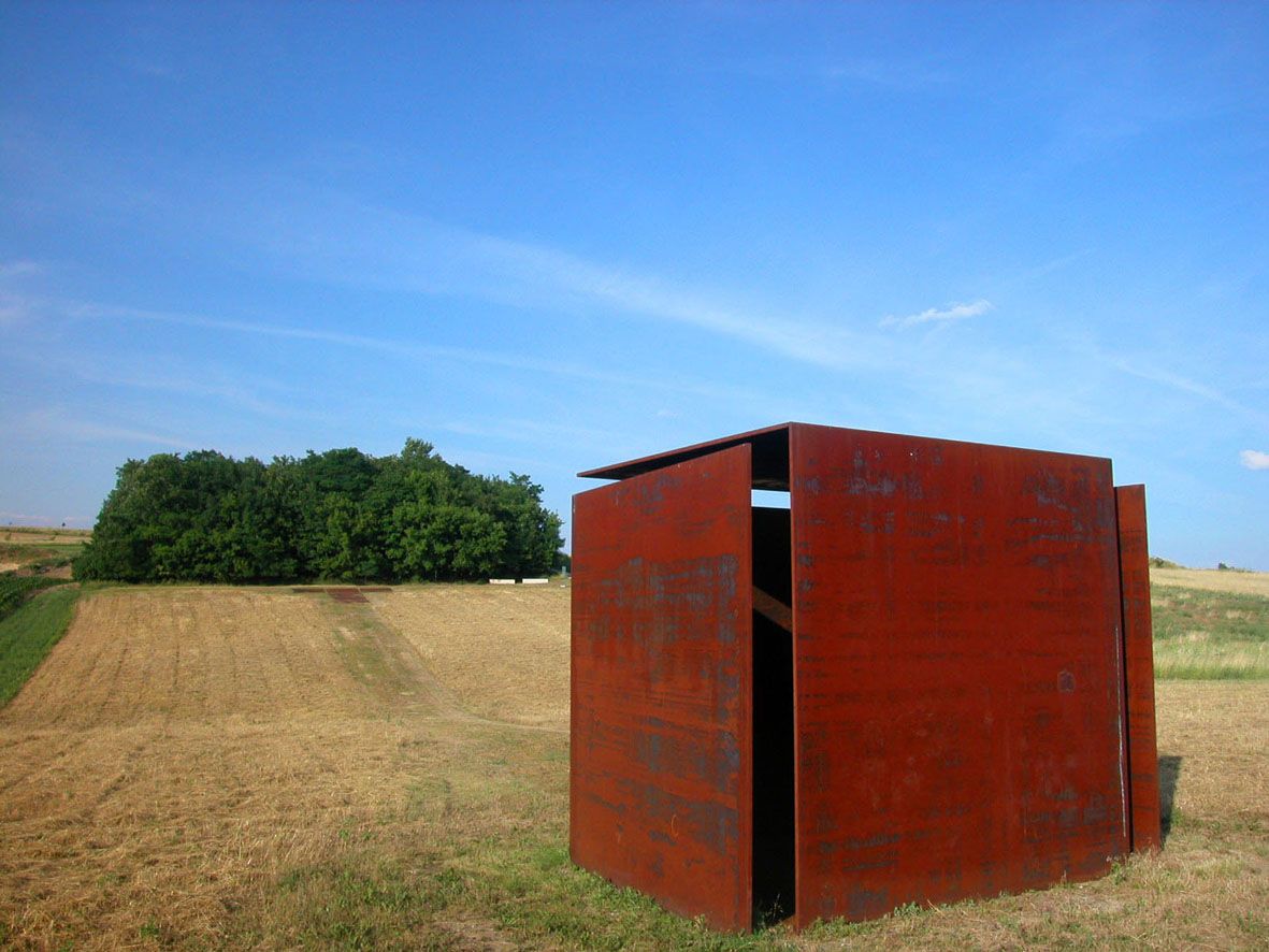 Eine rostige Metallskulptur steht auf einem Feld vor einem Wald unter blauem Himmel.