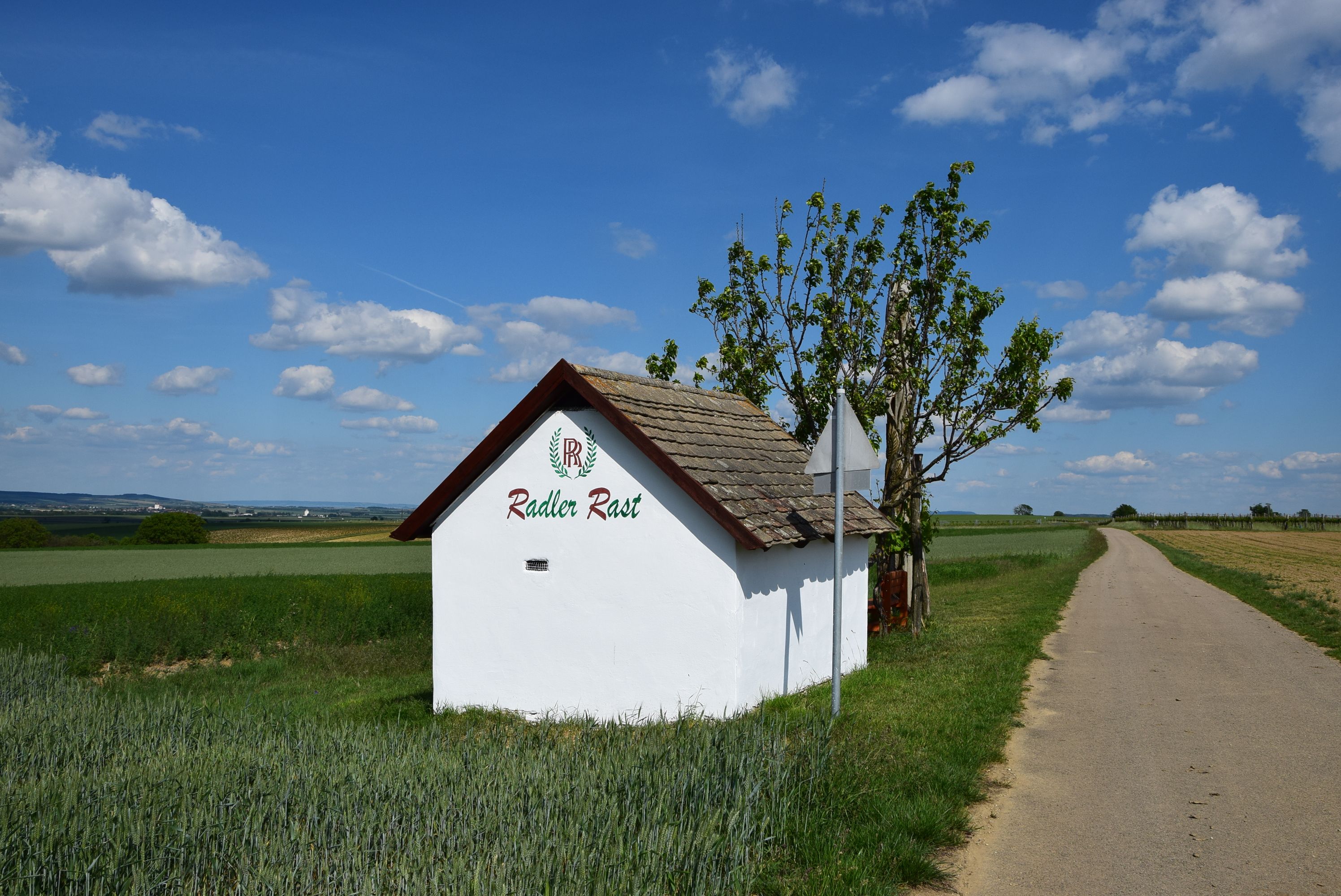 Kleines weißes Gebäude mit der Aufschrift 'Radler Rast' neben einem Feldweg unter blauem Himmel.