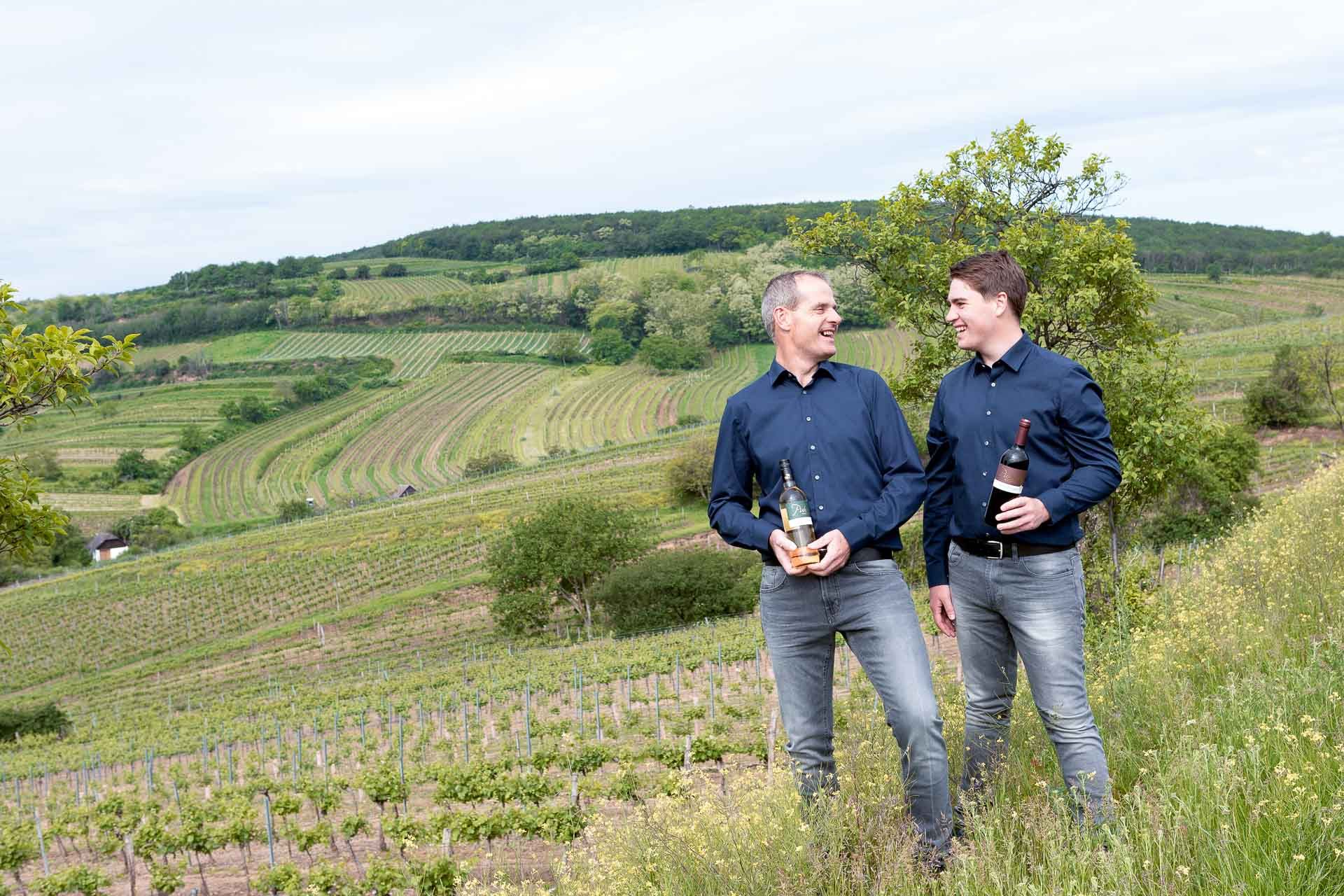 Two men in blue shirts are standing in a vineyard holding bottles of wine.