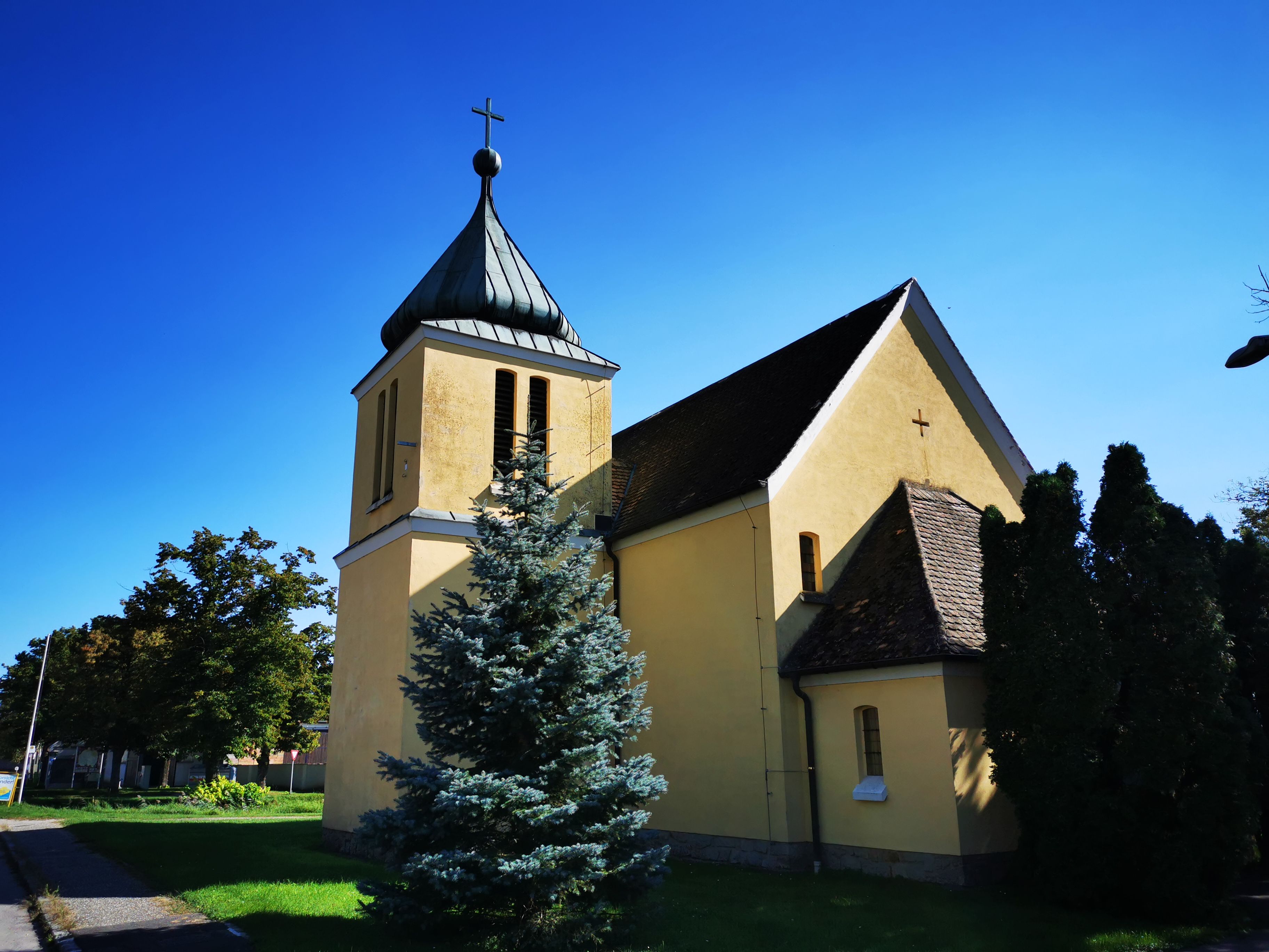 Žlutá kaple s věží a křížem, obklopená stromy, pod modrou oblohou.