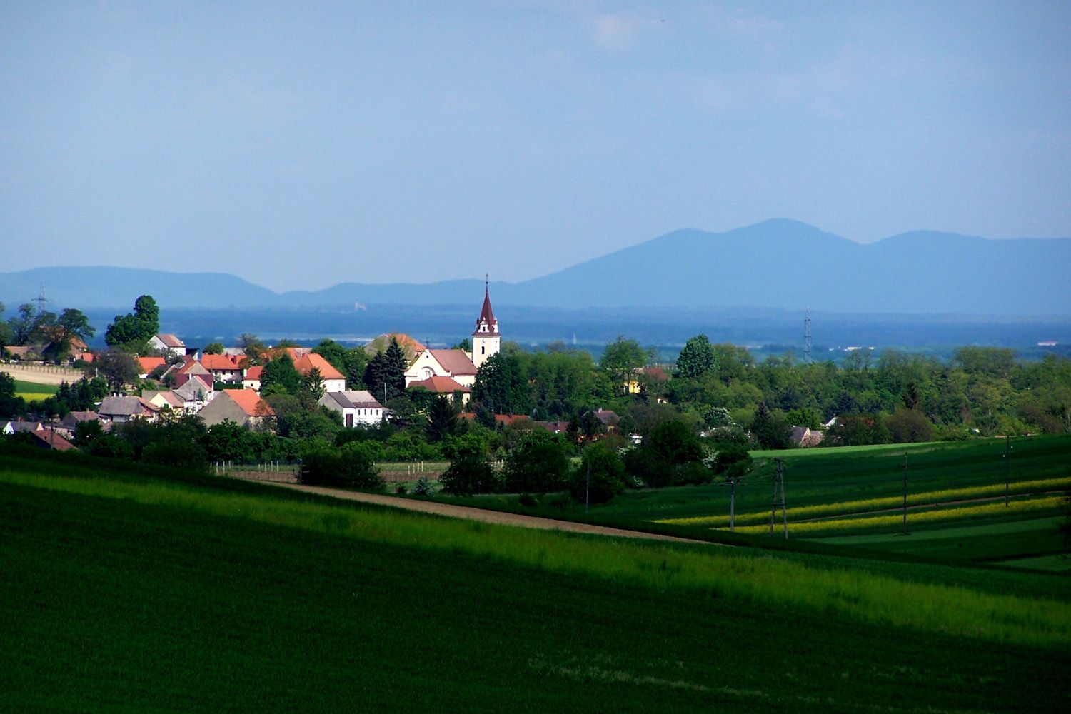 Landschaft mit Dorf und Kirche, umgeben von grünen Feldern und Hügeln im Hintergrund.