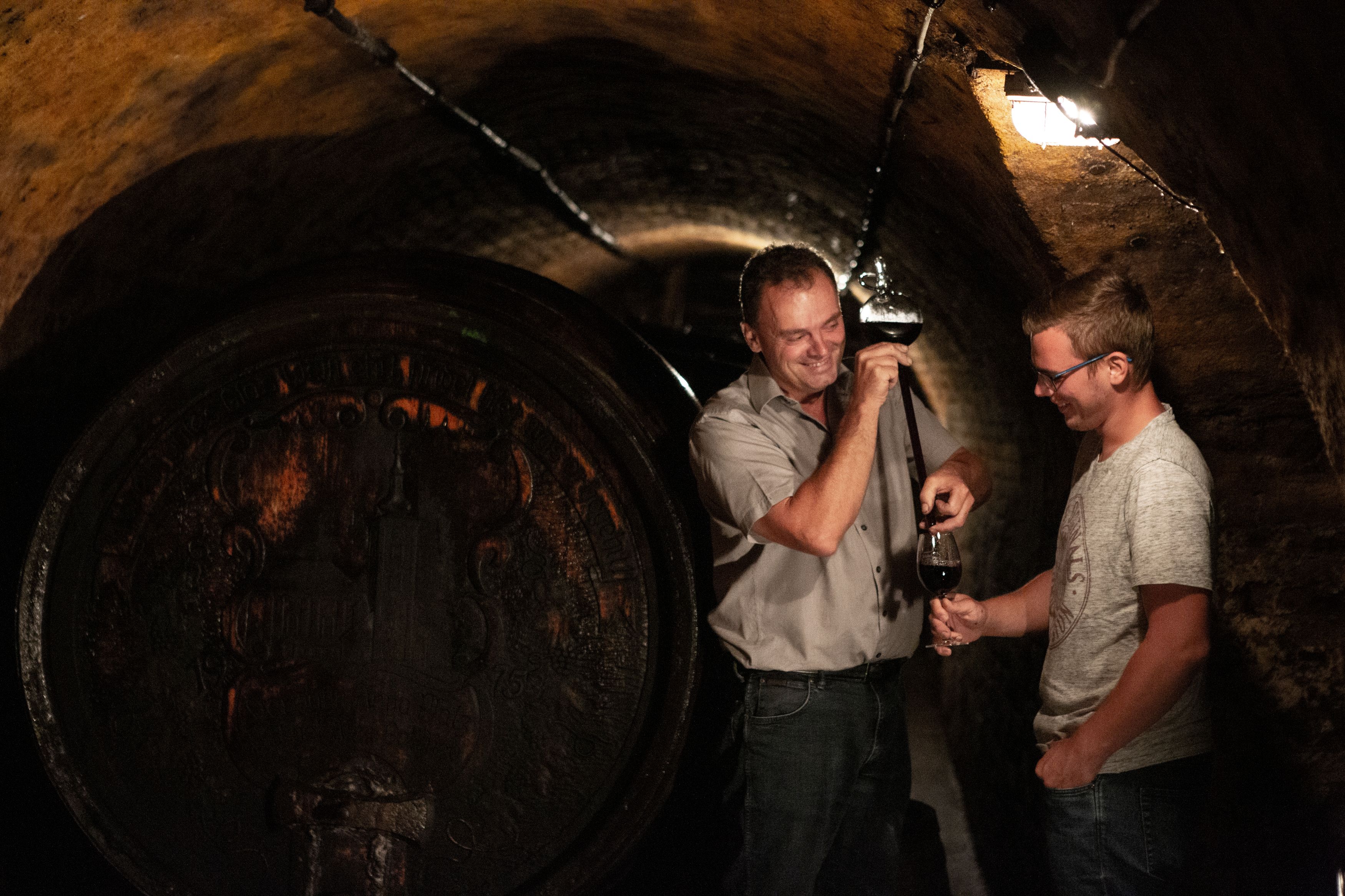 Two men in a wine cellar, one is holding a wine tasting from a barrel.
