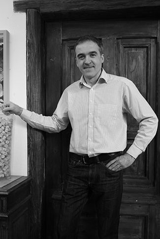 A man in a shirt stands in front of a wooden door and smiles at the camera.