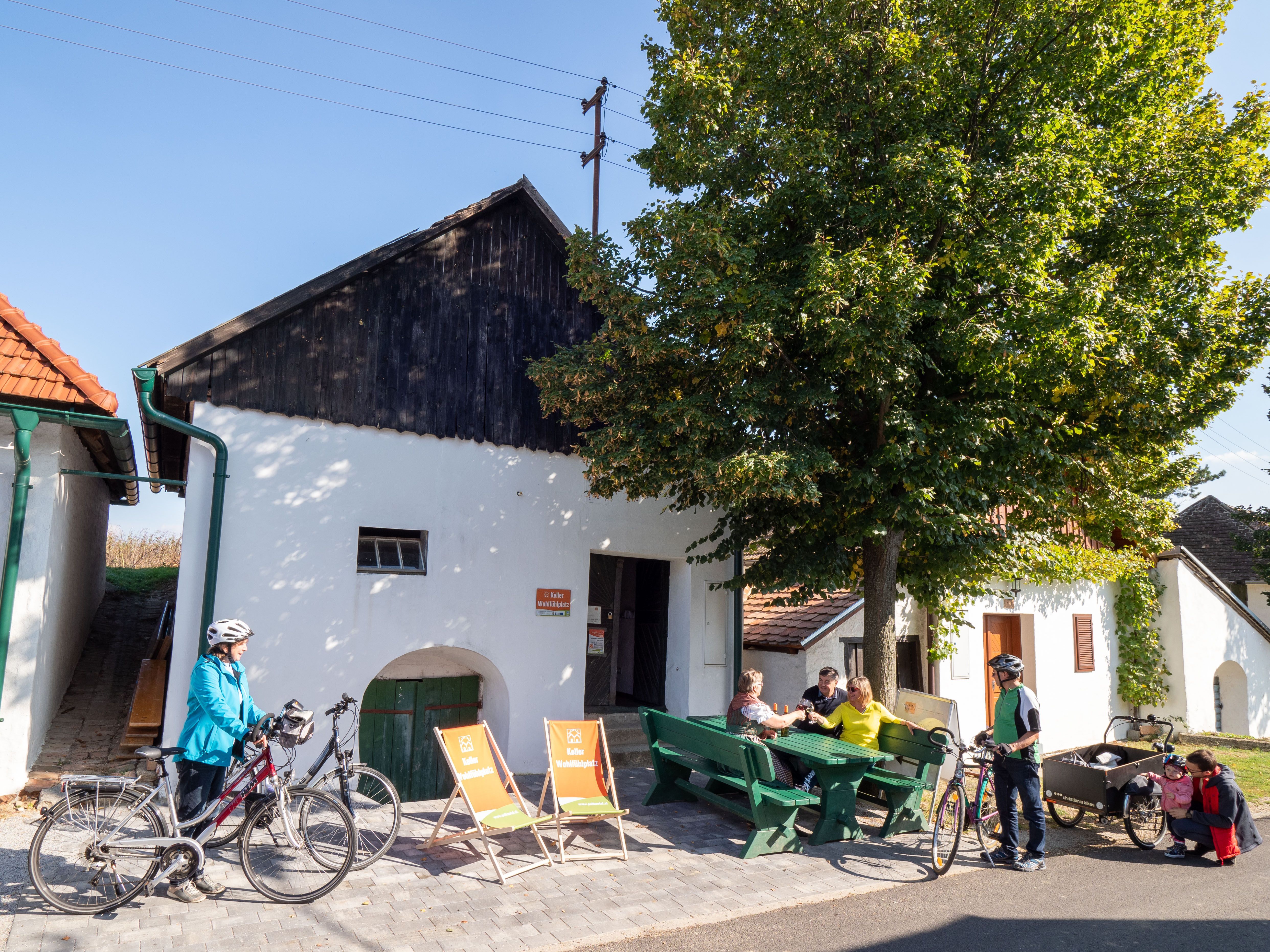 Ľudia sedia na lavičkách pred bielou budovou s bicyklami v Alberndorfe.
