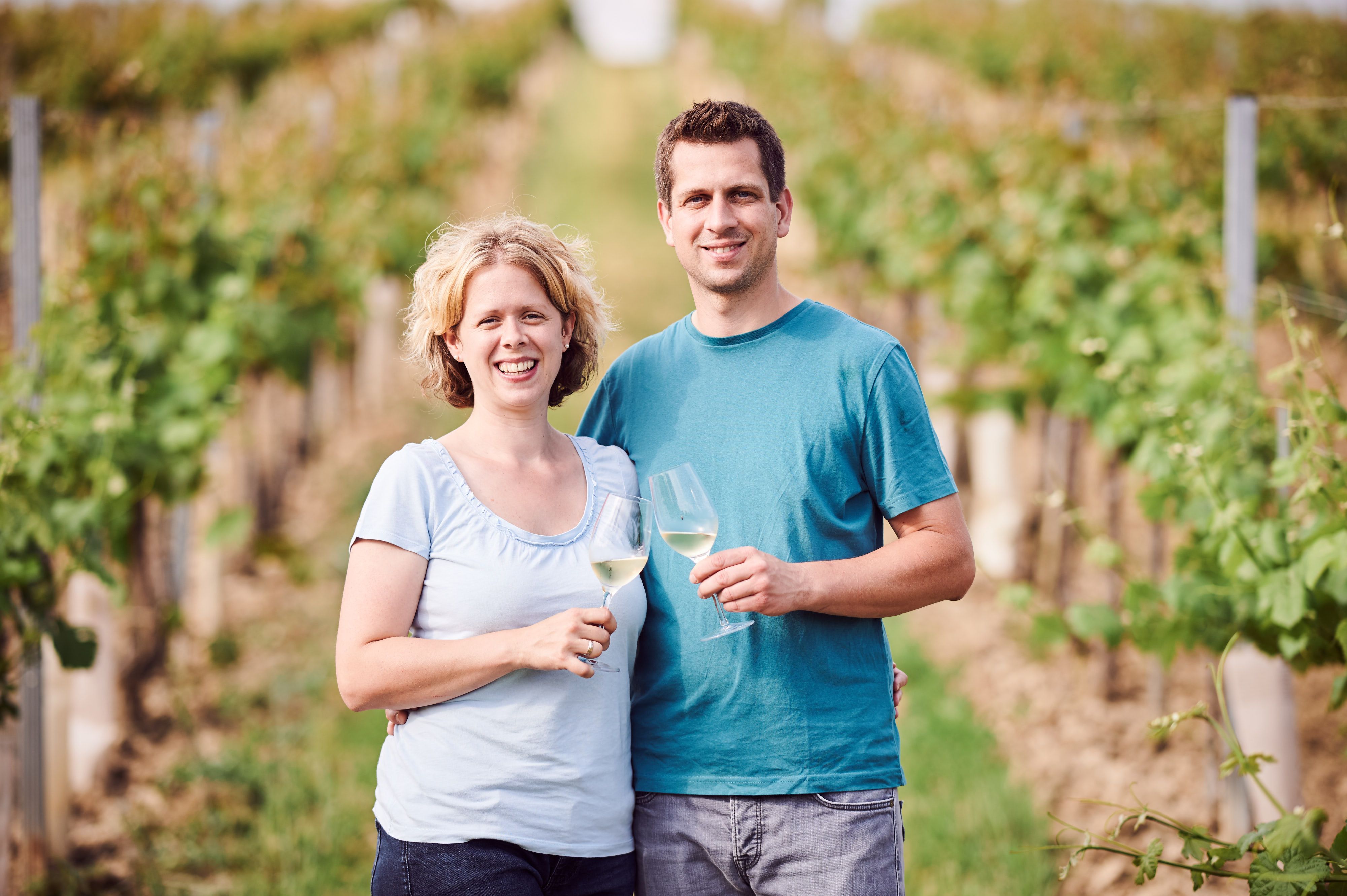 A couple stands in a vineyard holding glasses of wine.