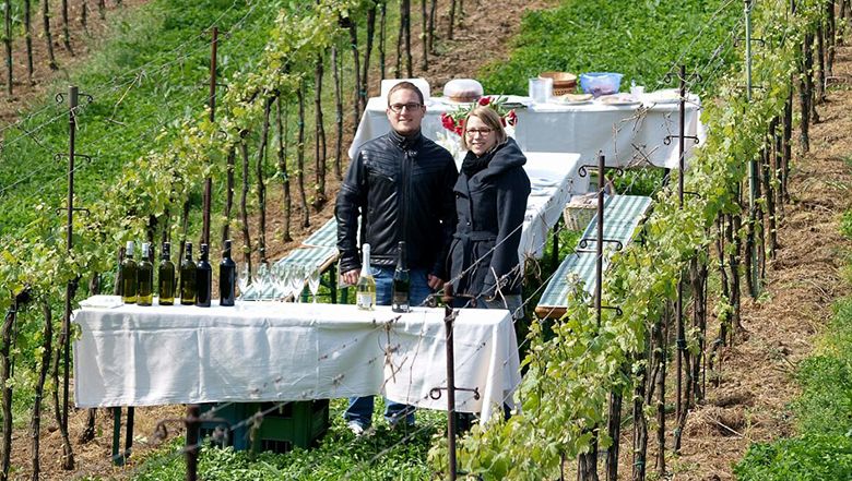 Two people are standing in a vineyard next to a laid table with bottles of wine.