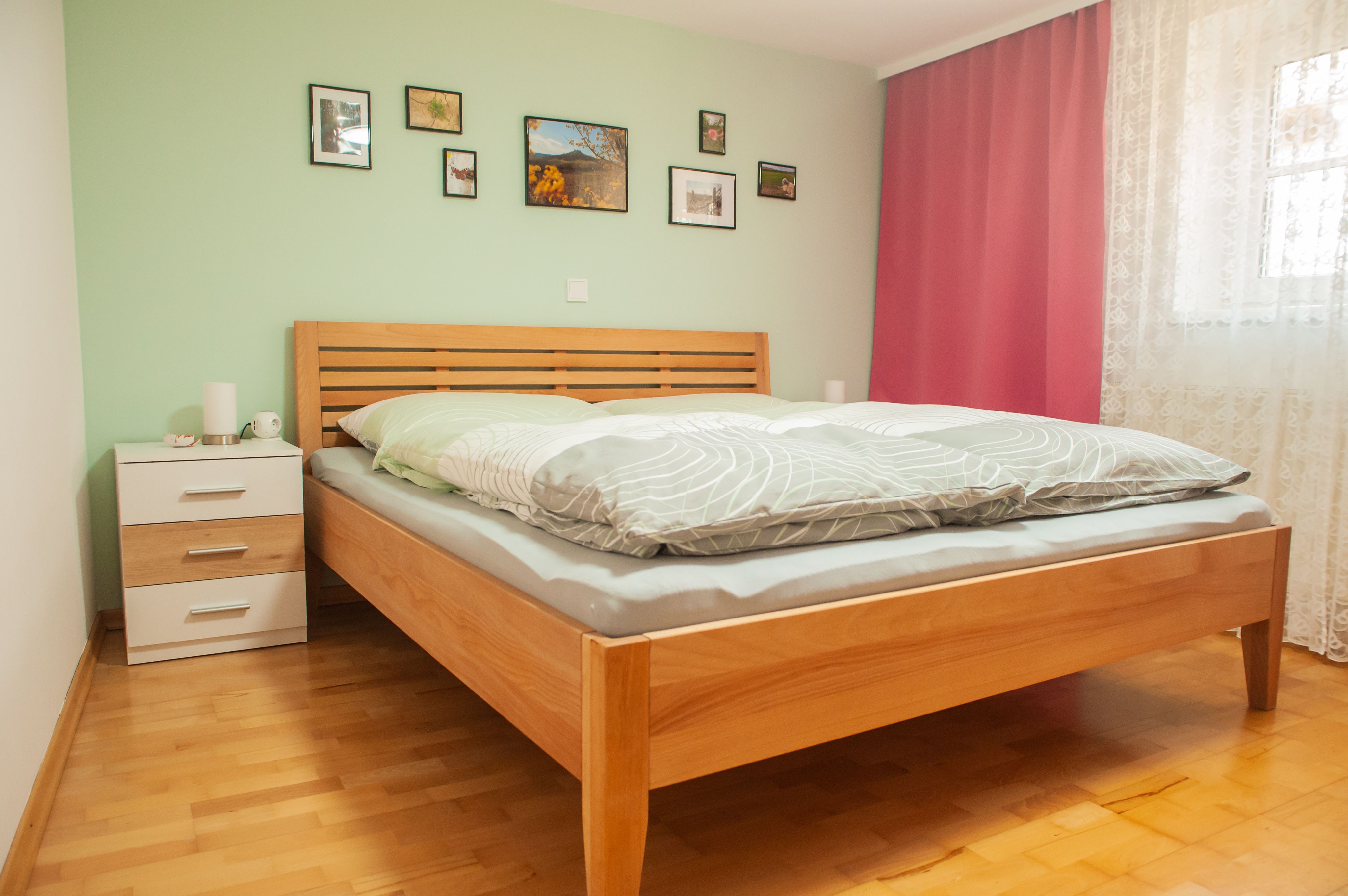 Spálňa s drevenou posteľou, zelenou stenou, obrázkami, nočným stolíkom a ružovými závesmi.